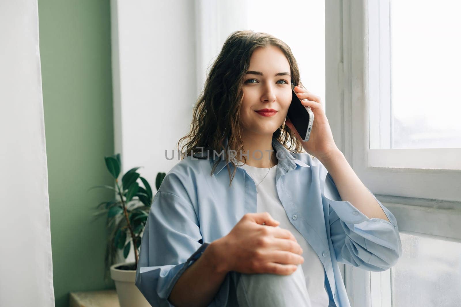A busy but happy mom enjoying a phone call with her best friend while kids play in the background. Casual indoor lifestyle portrait of a charming 30s woman sitting on windowsill.