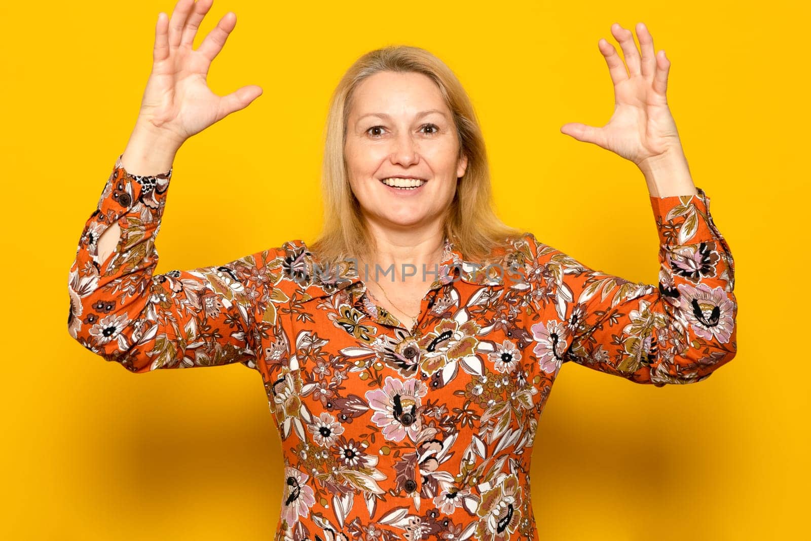 Blonde Caucasian woman in her 40s tremendously surprised by an unexpected visitor, raises her arms excitedly. Isolated on yellow studio background