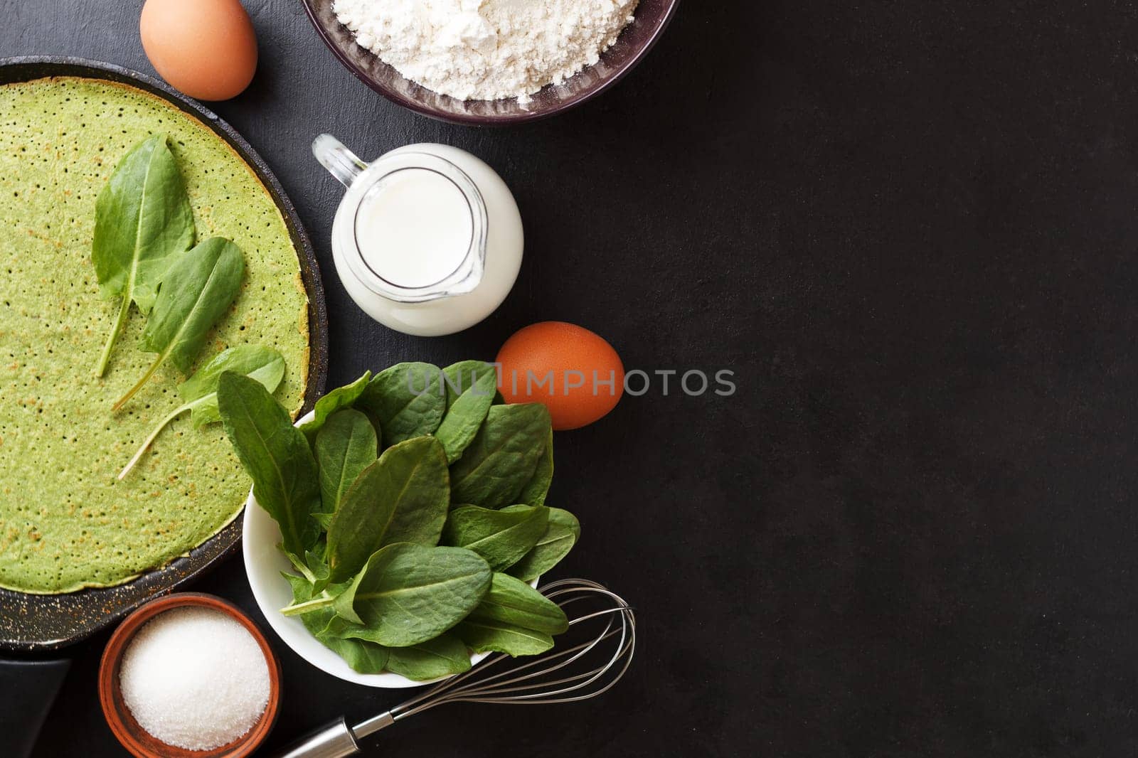 Spinach pancake in a pan on a black background with ingredients: eggs, spinach, milk, flour. Cooking. copy space