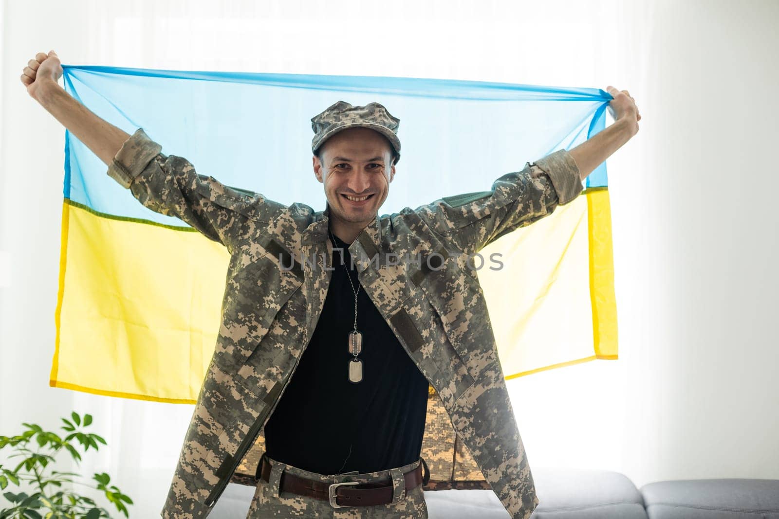 Ukrainian man warrior dressed in a military pixel uniform holds the yellow-blue flag of the state of Ukraine and on the chain a small coat of arms of the country of trident, close up. by Andelov13