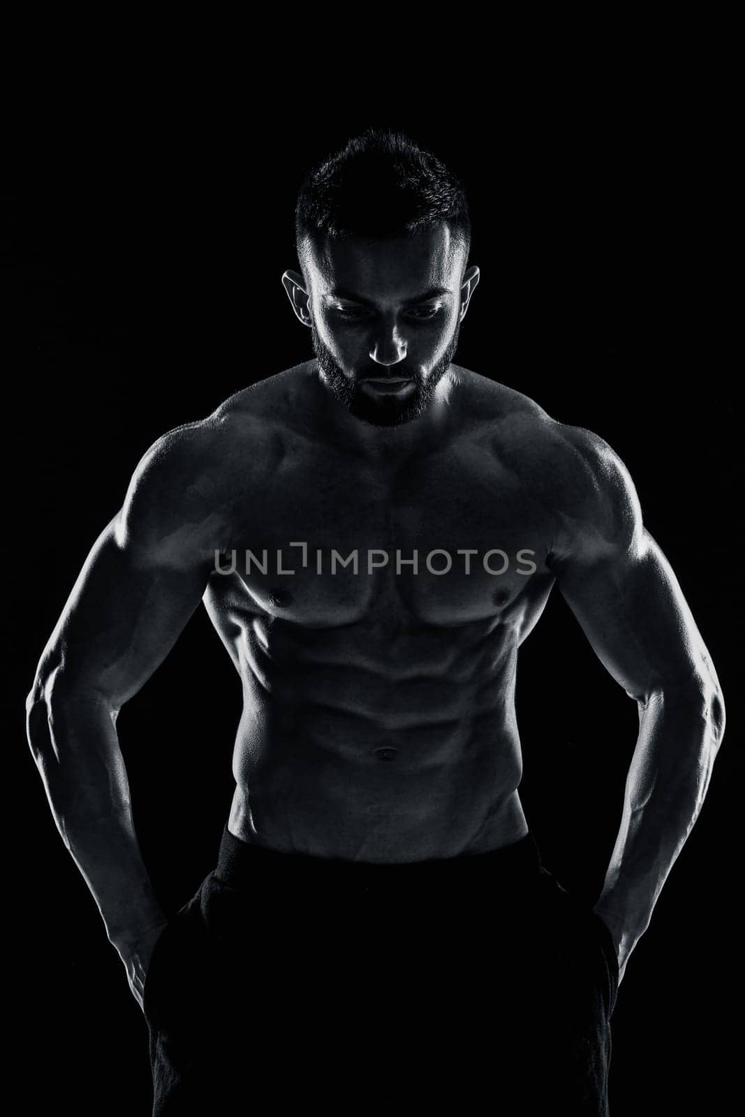 Image of very muscular man posing with naked torso in studio on black background. Black and white color