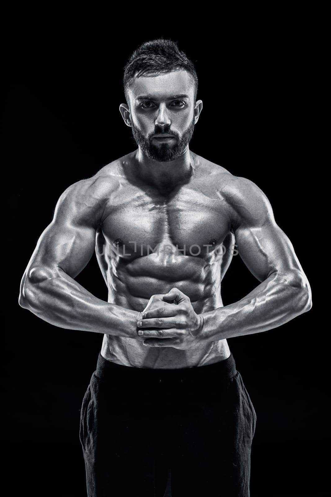 Image of very muscular man posing with naked torso in studio on black background. Black and white color