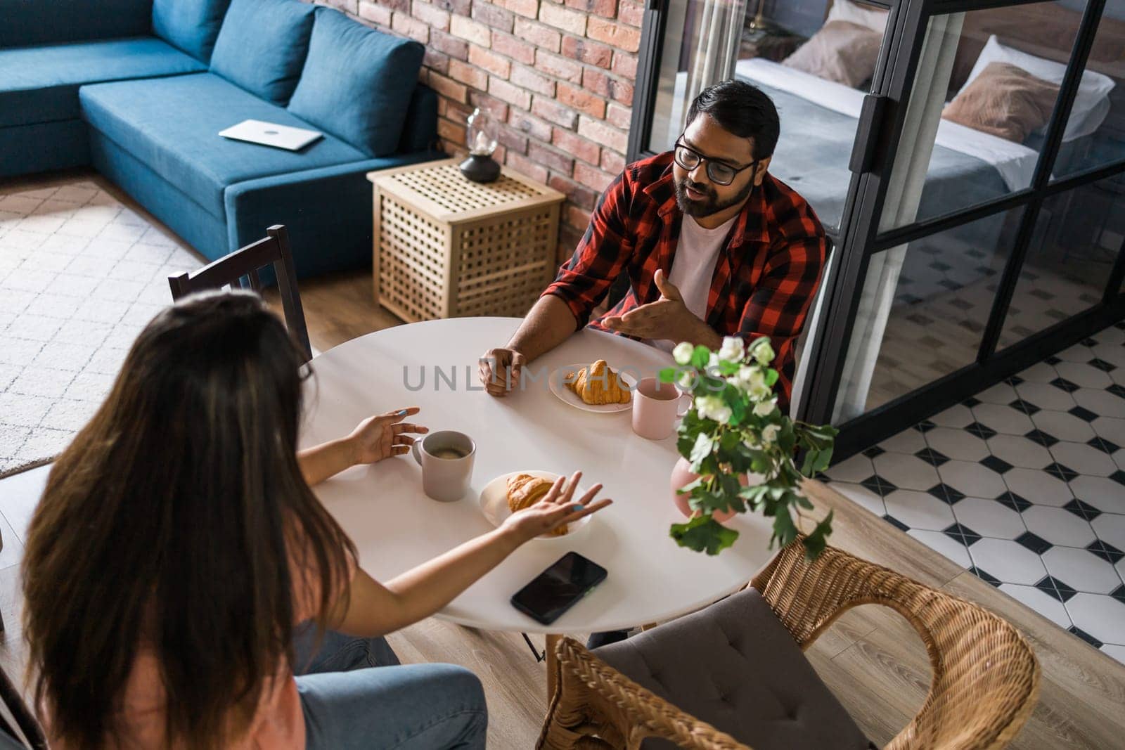 Young diverse loving couple eating croissant and talks together at home in breakfast time. Communication and relationship