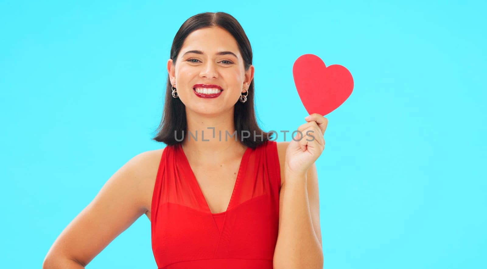 Paper heart, happy woman and face on blue background, studio and backdrop. Portrait of female model in red dress with shape of love, trust and romance for valentines day, flirting and elegant smile.