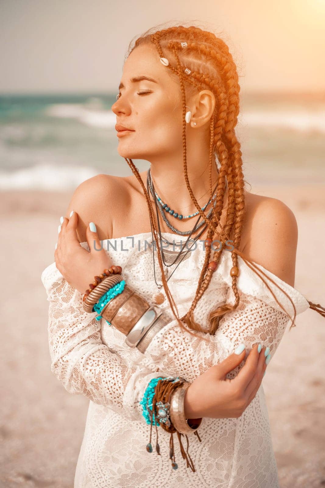 Model in boho style in a white long dress and silver jewelry on the beach. Her hair is braided, and there are many bracelets on her arms. by Matiunina