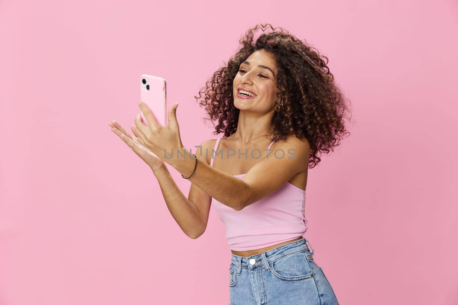 Woman blogger holding phone in hand video call, with curly hair in pink top and jeans poses on pink background, copy space, technology and social media, online. High quality photo