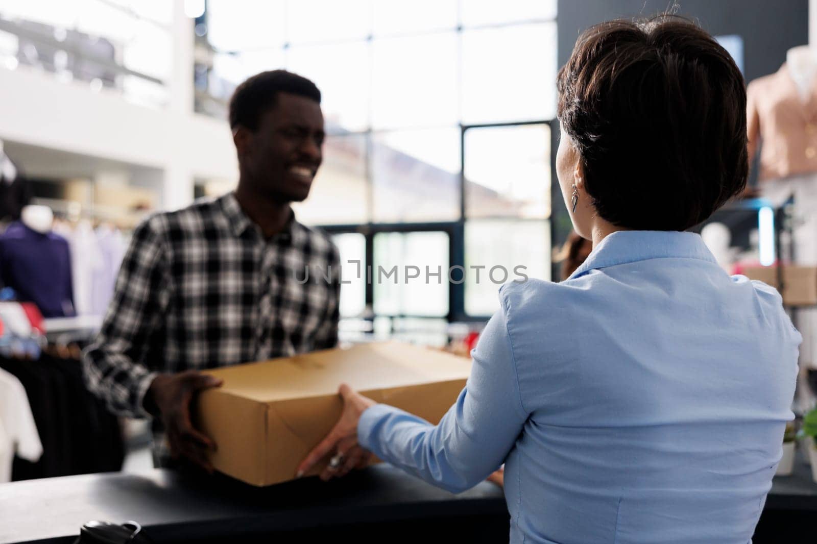 African american man standing at store counter desk, holding carton box with stylish clothes in modern boutique. Customer discussing order price with fashionable employee. Fashion concept