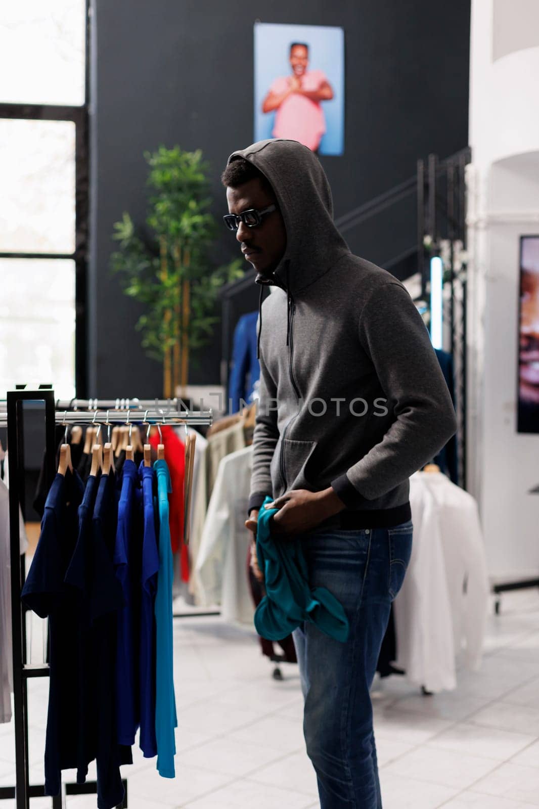 African american man thief looking at trendy clothes before stealing it in modern boutique. Robber wearing sunglasses and hood not be recognized in clothing store. Burglary concept