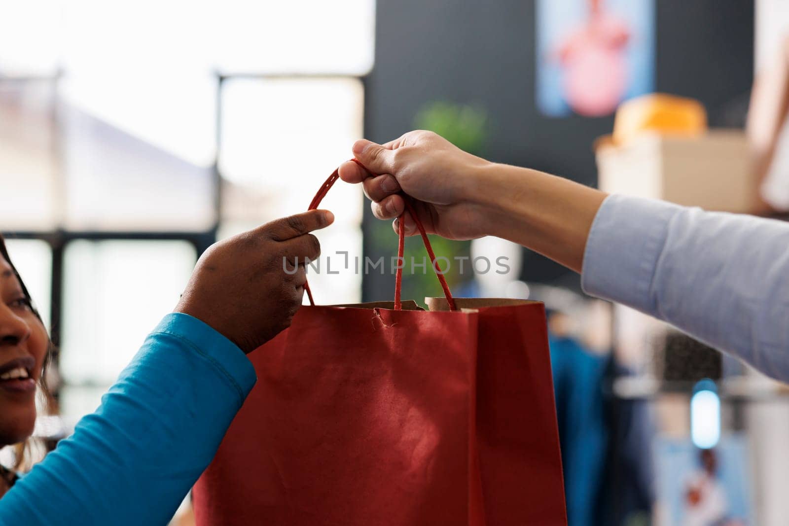 Employee giving red paper bag to customer after purchasing fashionable clothes for new wardrobe in modern boutique. African american woman shopping for casual wear in clothing store