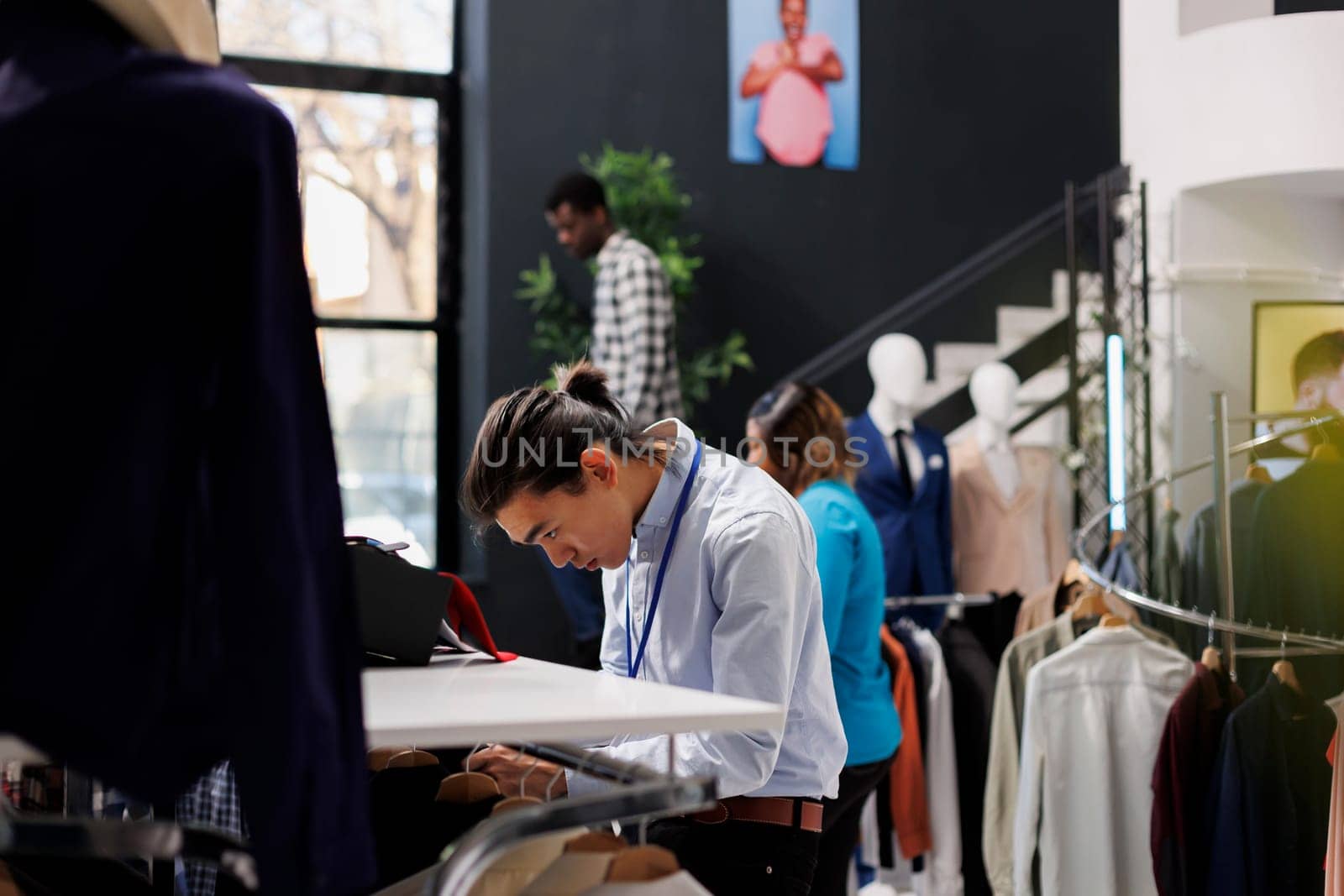 Asian shopping mall worker checking clothes, putting fashionable merchandise on hangers, working at modern boutique visual. Stylish man arranging formal wear items in clothing store. Fashion concept