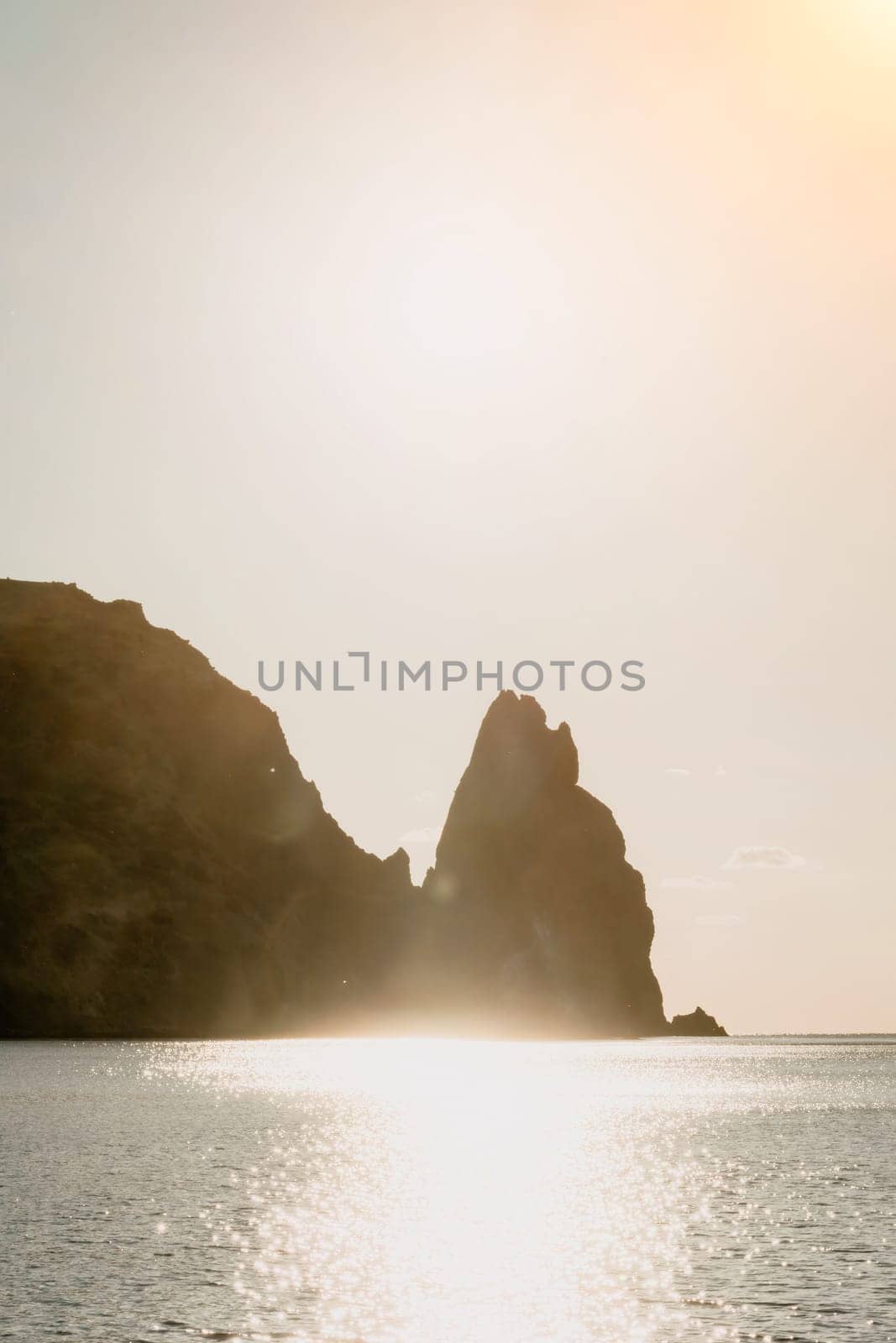 A red burning sunset with the silhouette of a cliff over the sea. Abstract nature summer or spring ocean sea background. Small waves on golden warm water surface with bokeh lights from sun.