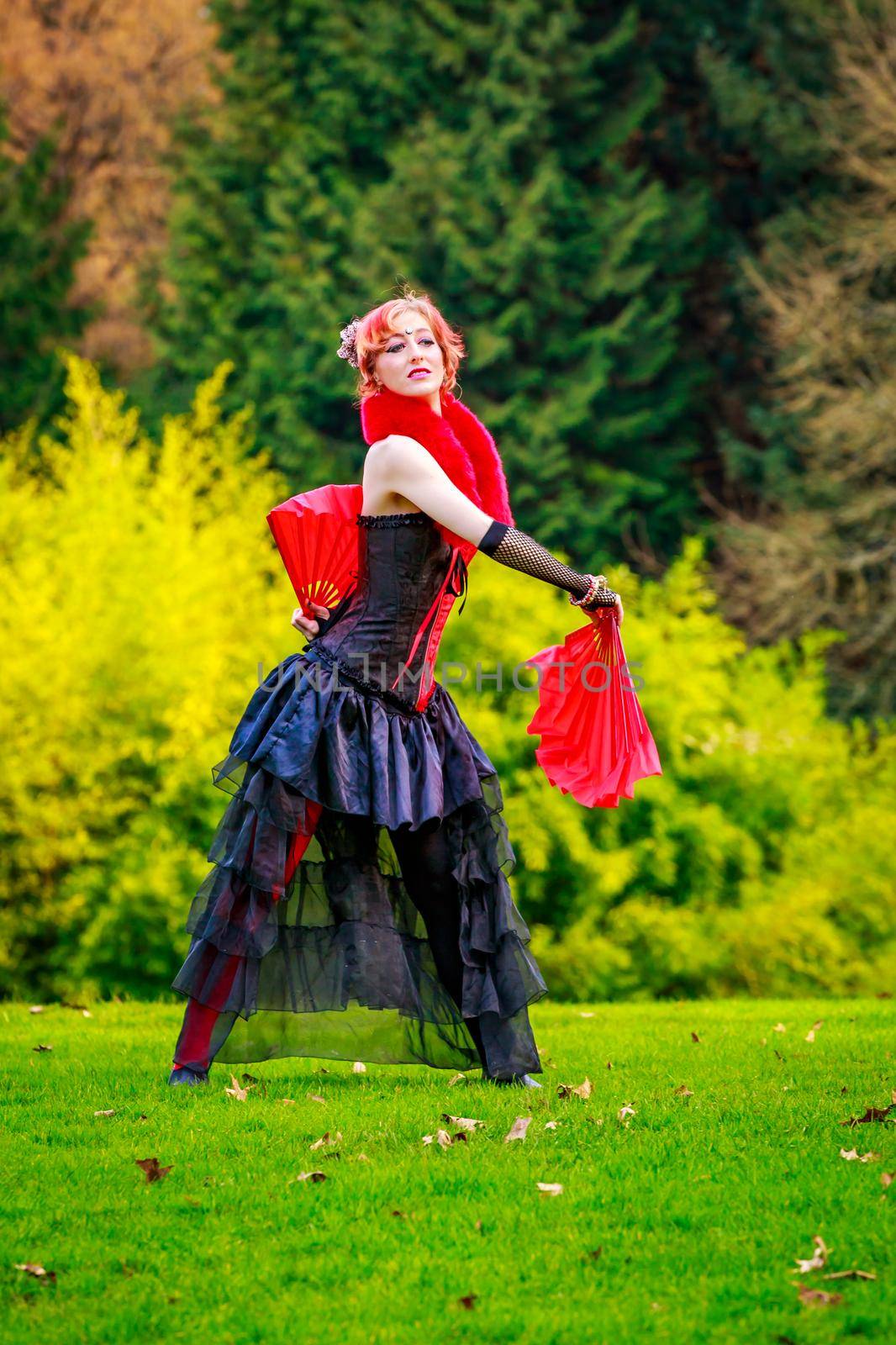 Young beautiful woman in circus costume play with red fans in the park.