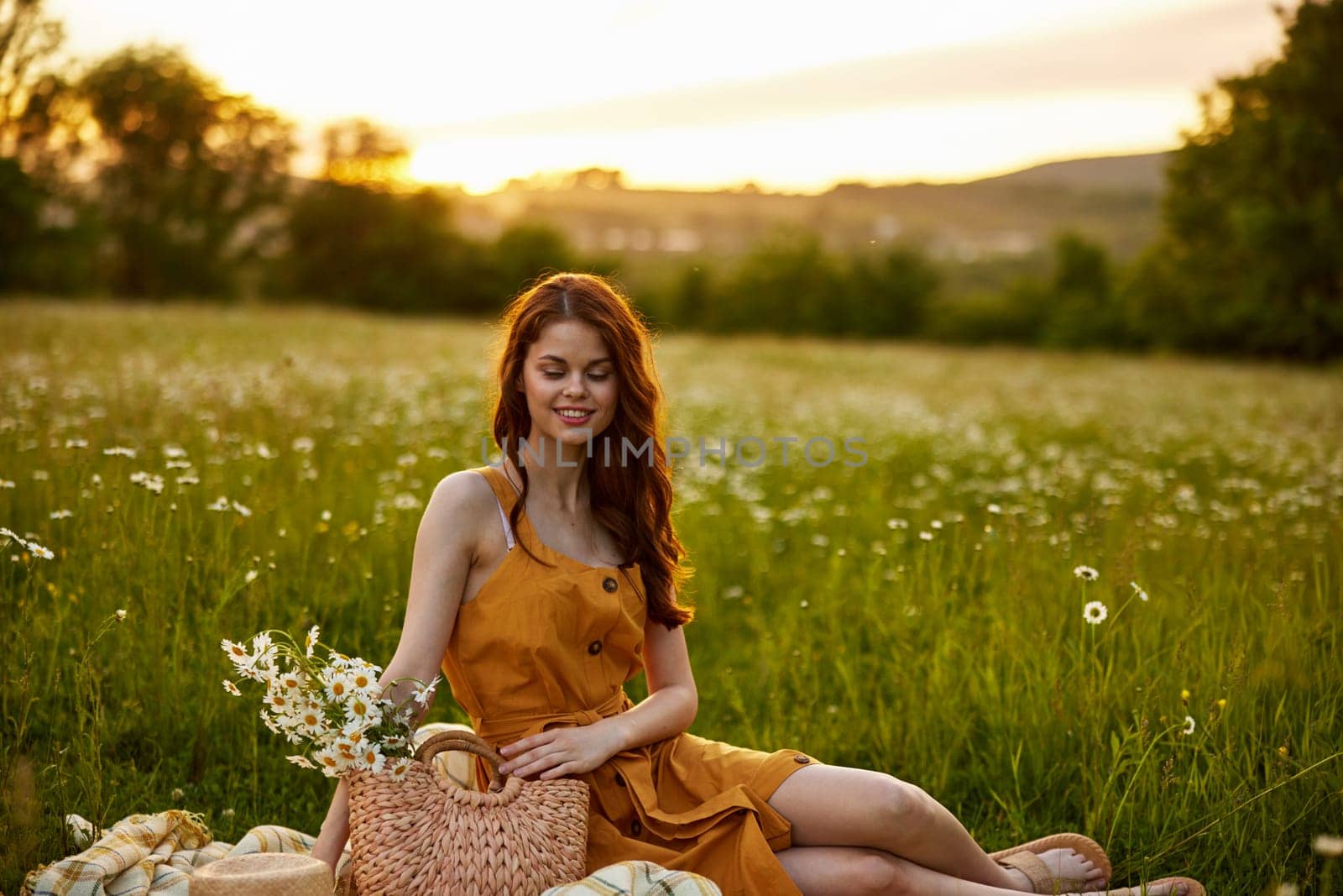 a happy woman in an orange dress sits on a plaid in a chamomile field at sunset and laughs. High quality photo