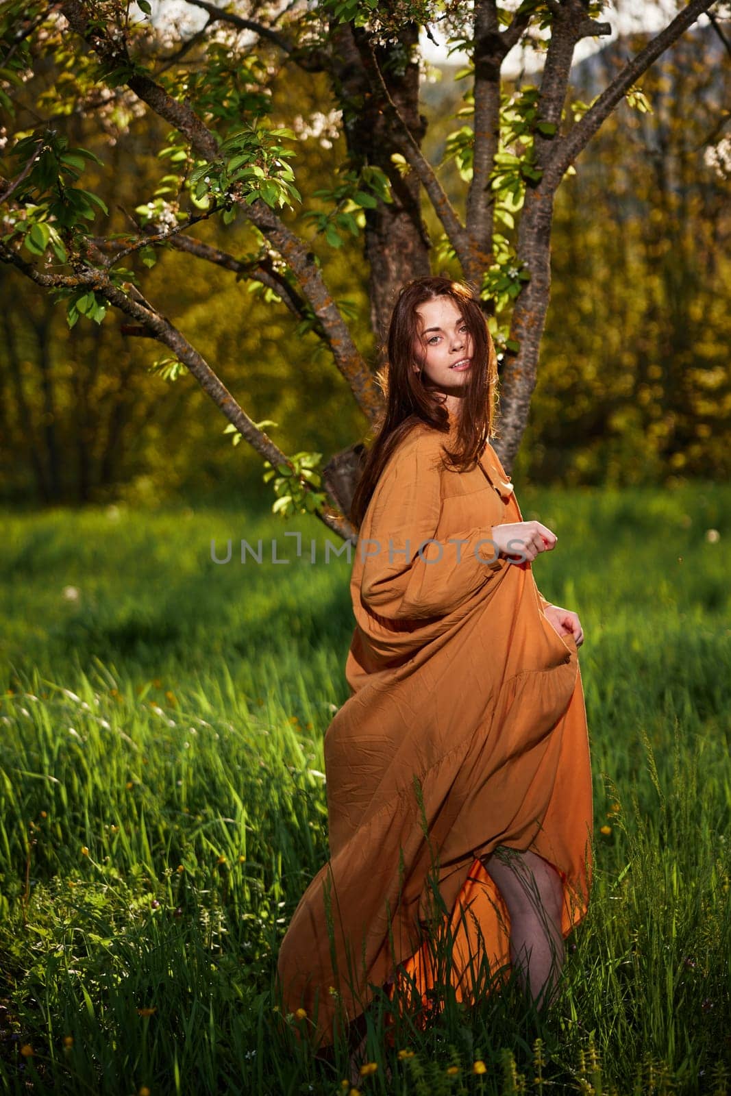 a sweet, modest, attractive woman with long red hair stands in the countryside near a flowering tree and lifts the hem of her dress up. High quality photo