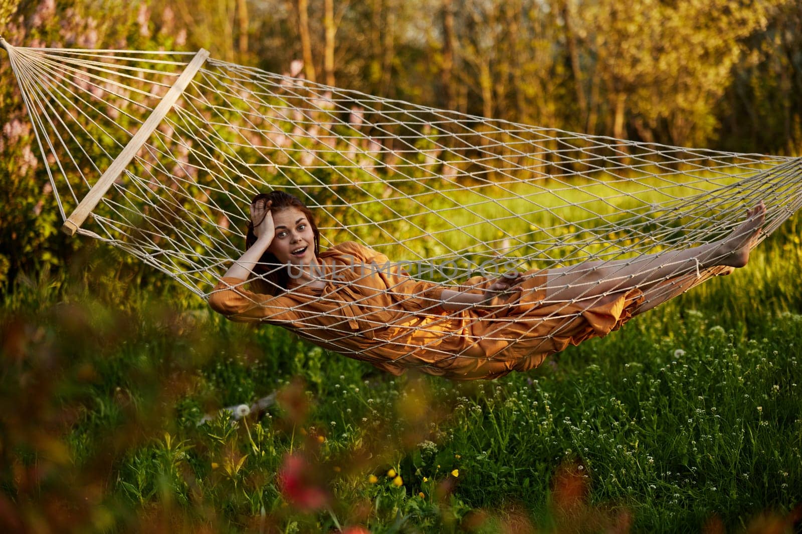 a beautiful woman is resting in nature lying in a mesh hammock in a long orange dress looking at the camera. Horizontal photo on the theme of recreation by Vichizh
