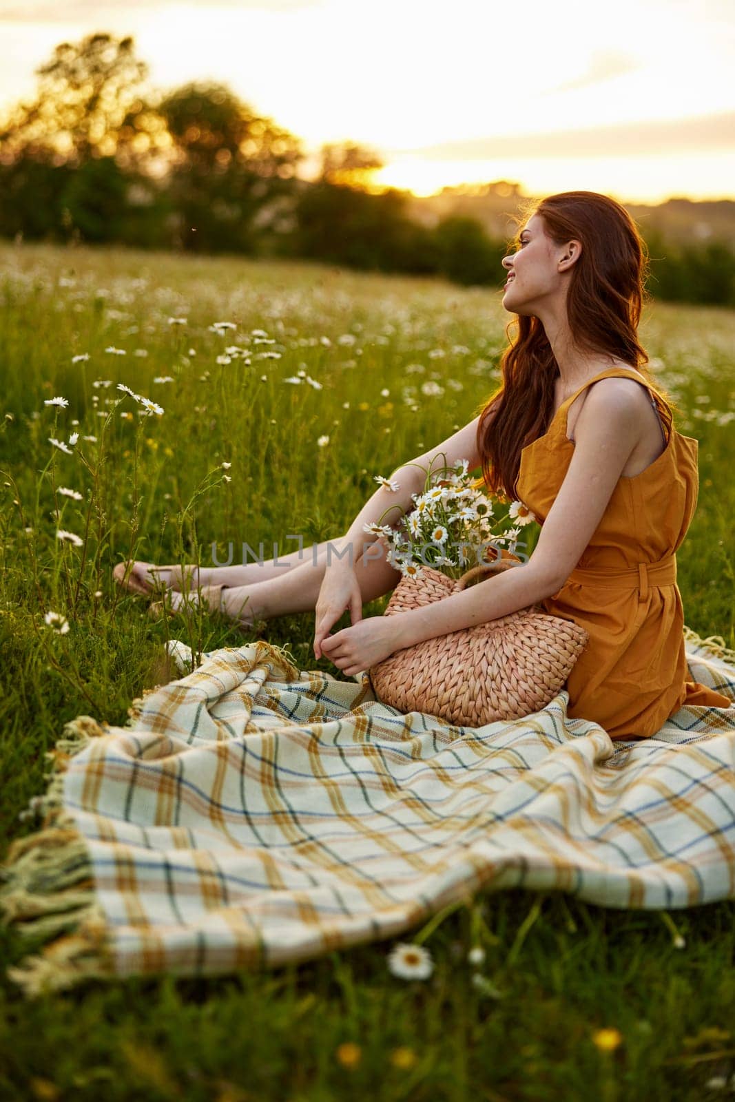 happy redhead woman sits in a field of daisies on a plaid during sunset and enjoys nature. High quality photo