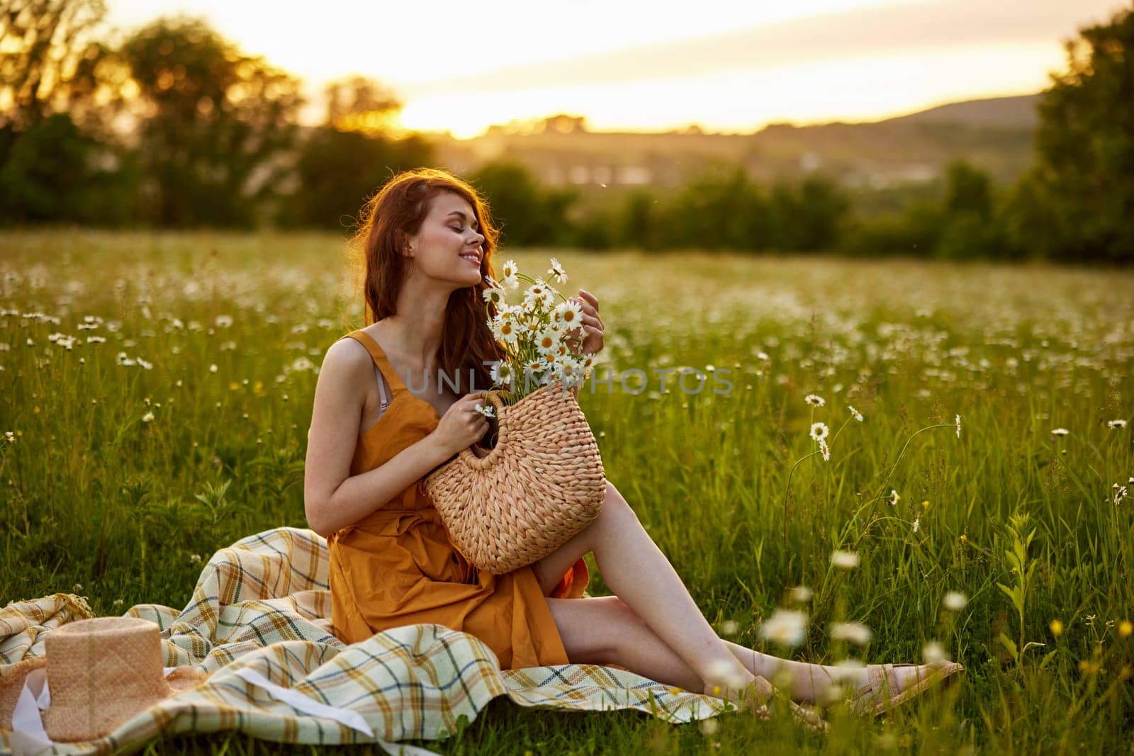 a happy woman in an orange dress sits on a plaid in a chamomile field at sunset and laughs. High quality photo