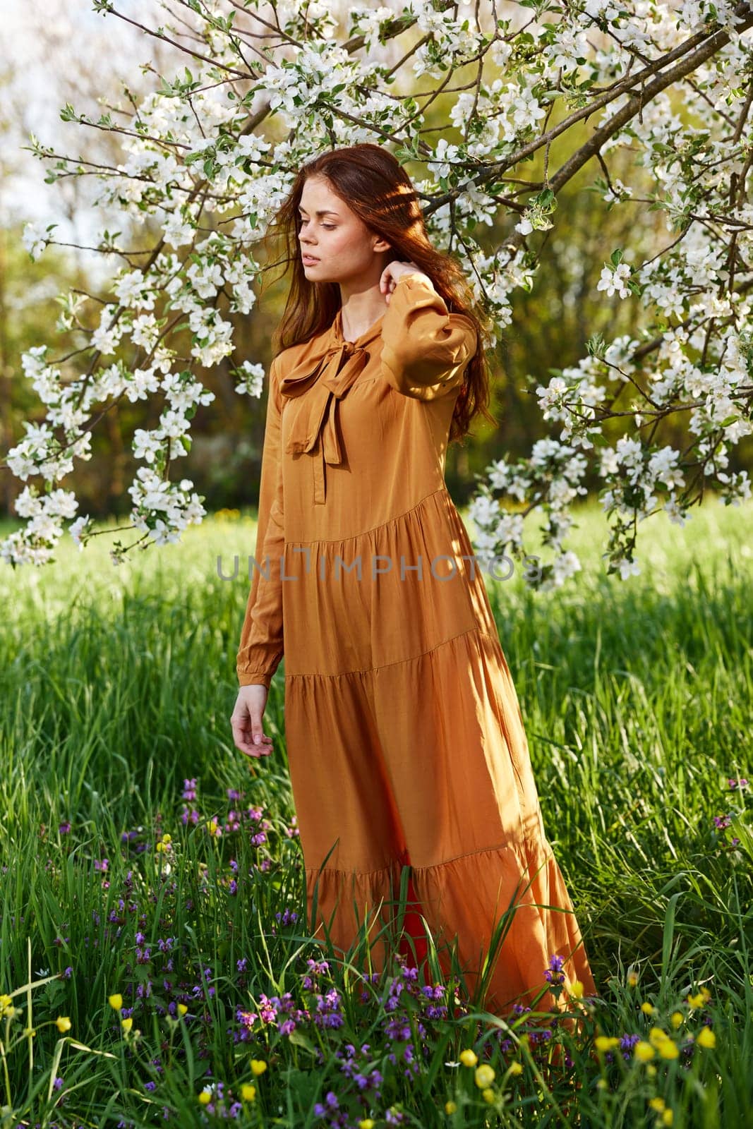 a bright vertical photo of an attractive woman in a long orange dress standing next to a flowering tree in sunny, warm weather, straightening her hair with her hand and closing her eyes. High quality photo