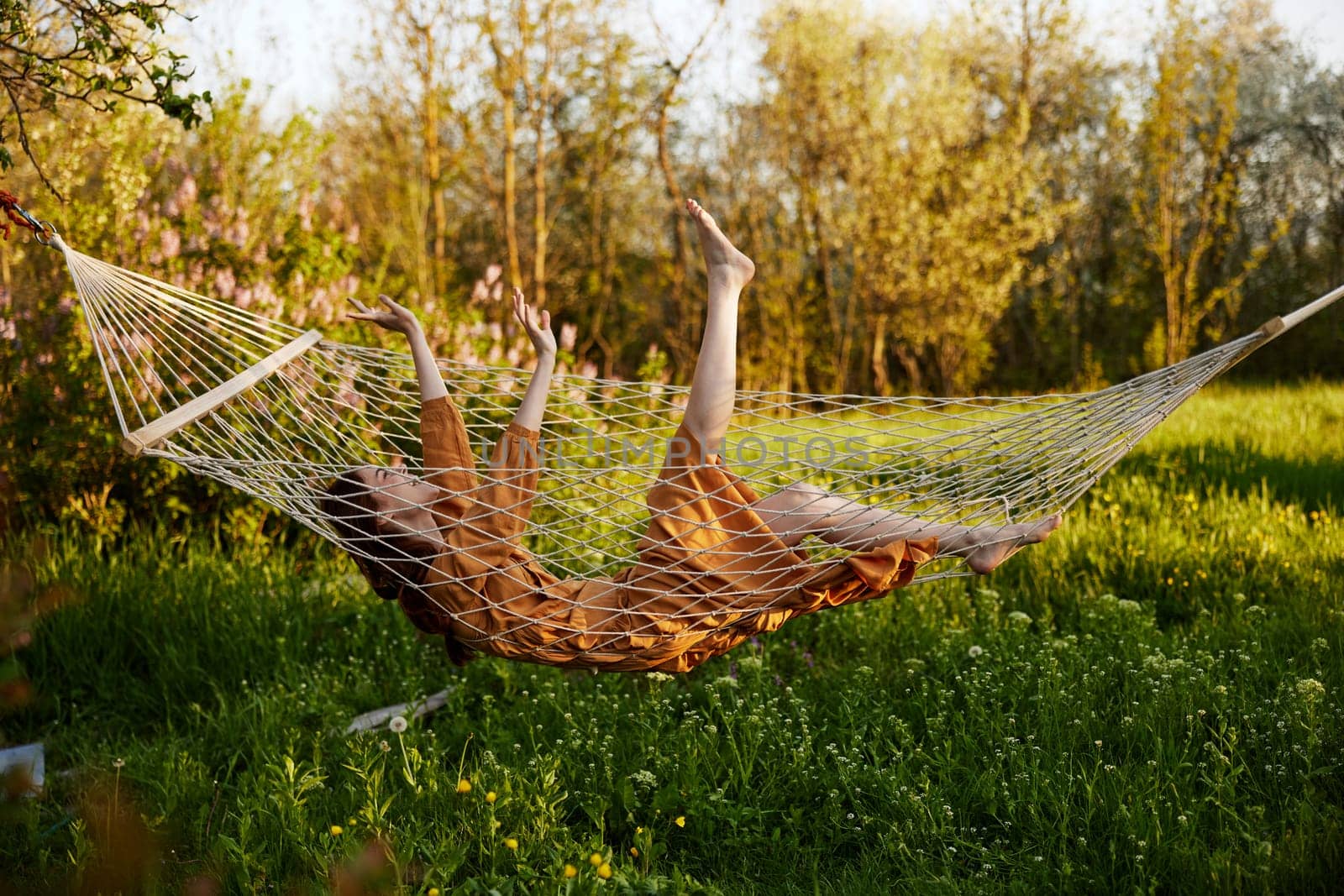 a funny woman is resting in nature lying in a mesh hammock in a long orange dress lifting up her arms and legs. High quality photo