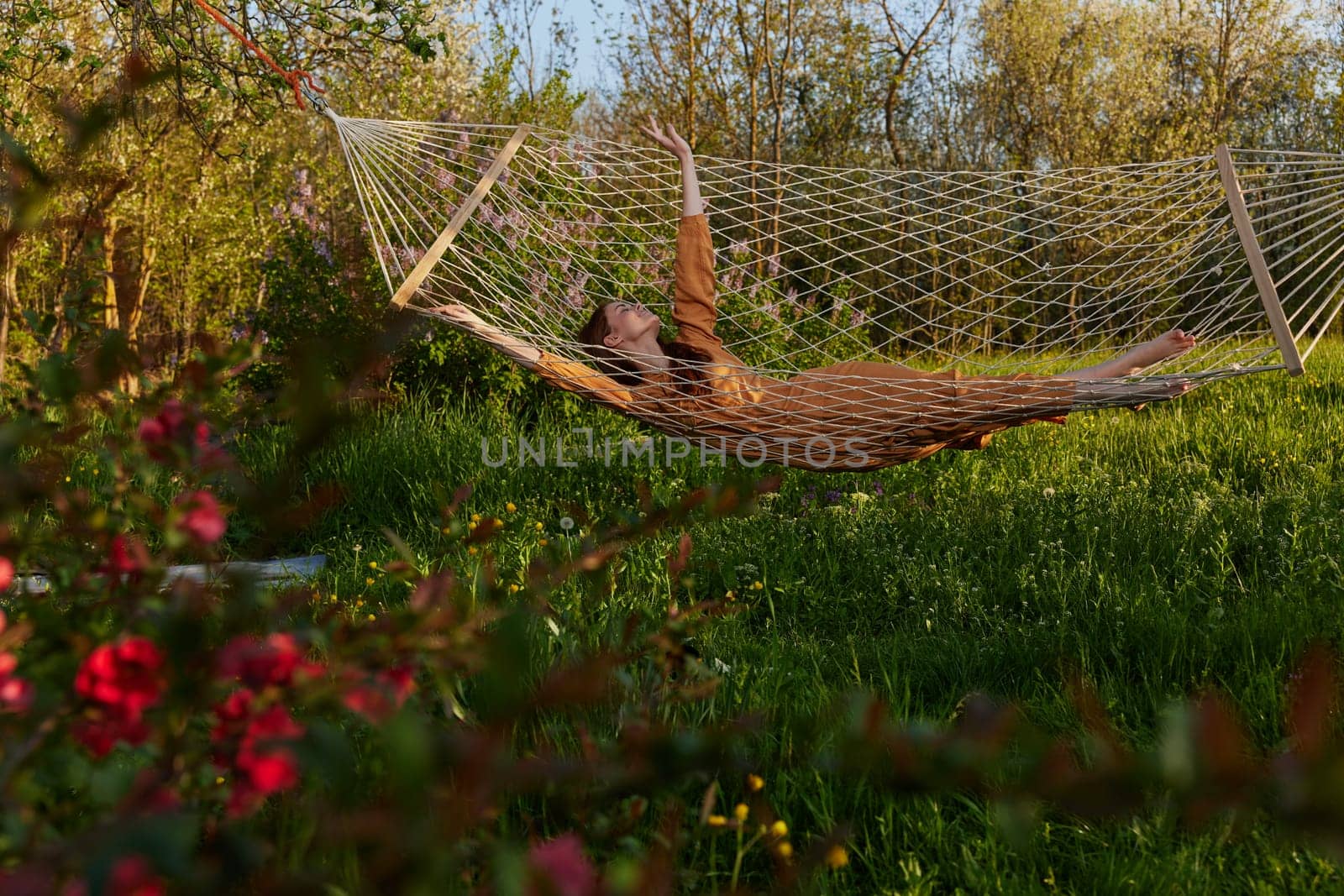 a happy woman in a long orange dress is relaxing in nature lying in a mesh hammock enjoying summer and vacation in the country surrounded by green foliage, happily lifting her legs. High quality photo