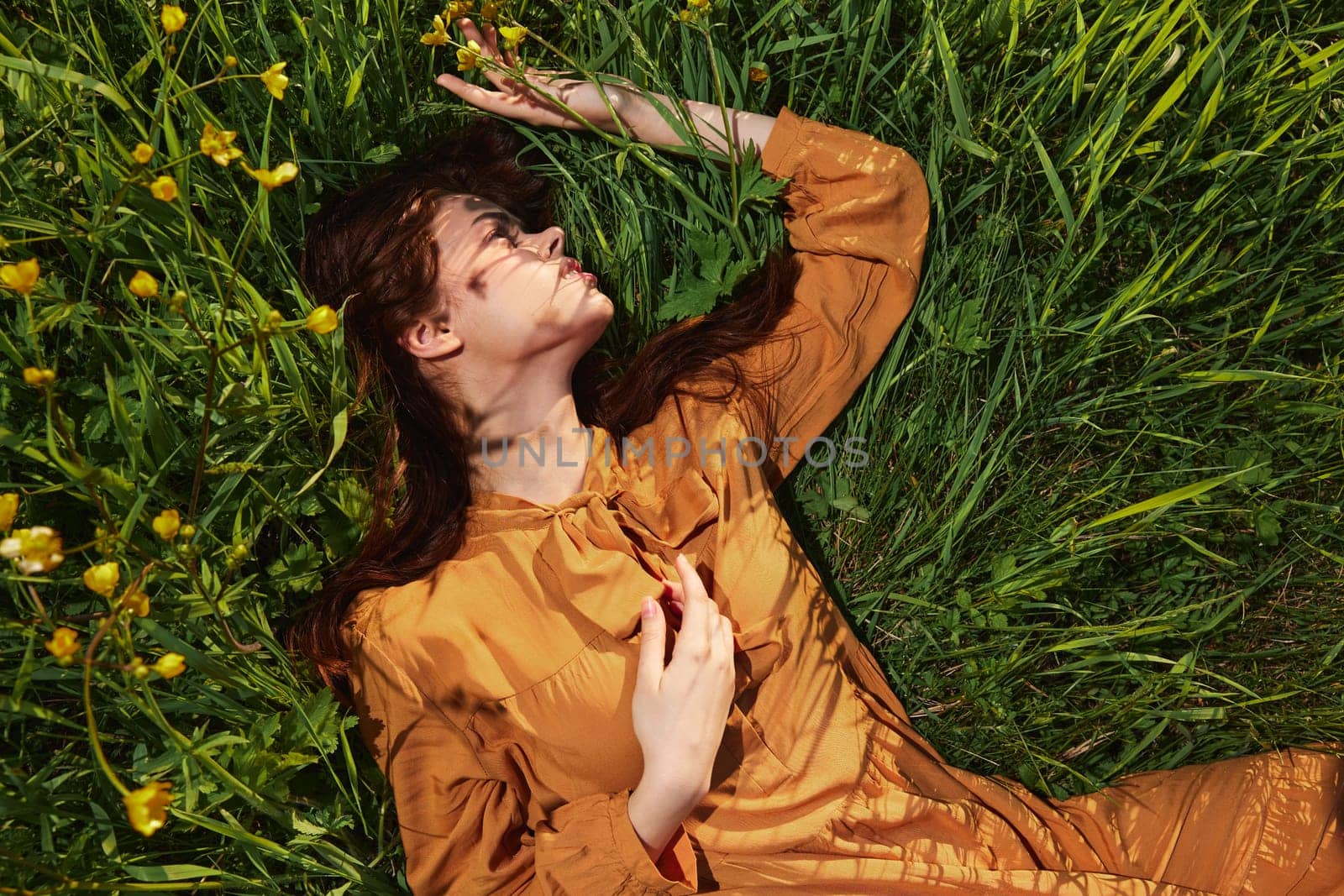 a calm woman with long red hair lies in a green field with yellow flowers, in an orange dress with her eyes closed, holding her hand near her face, enjoying peace and recuperating by Vichizh