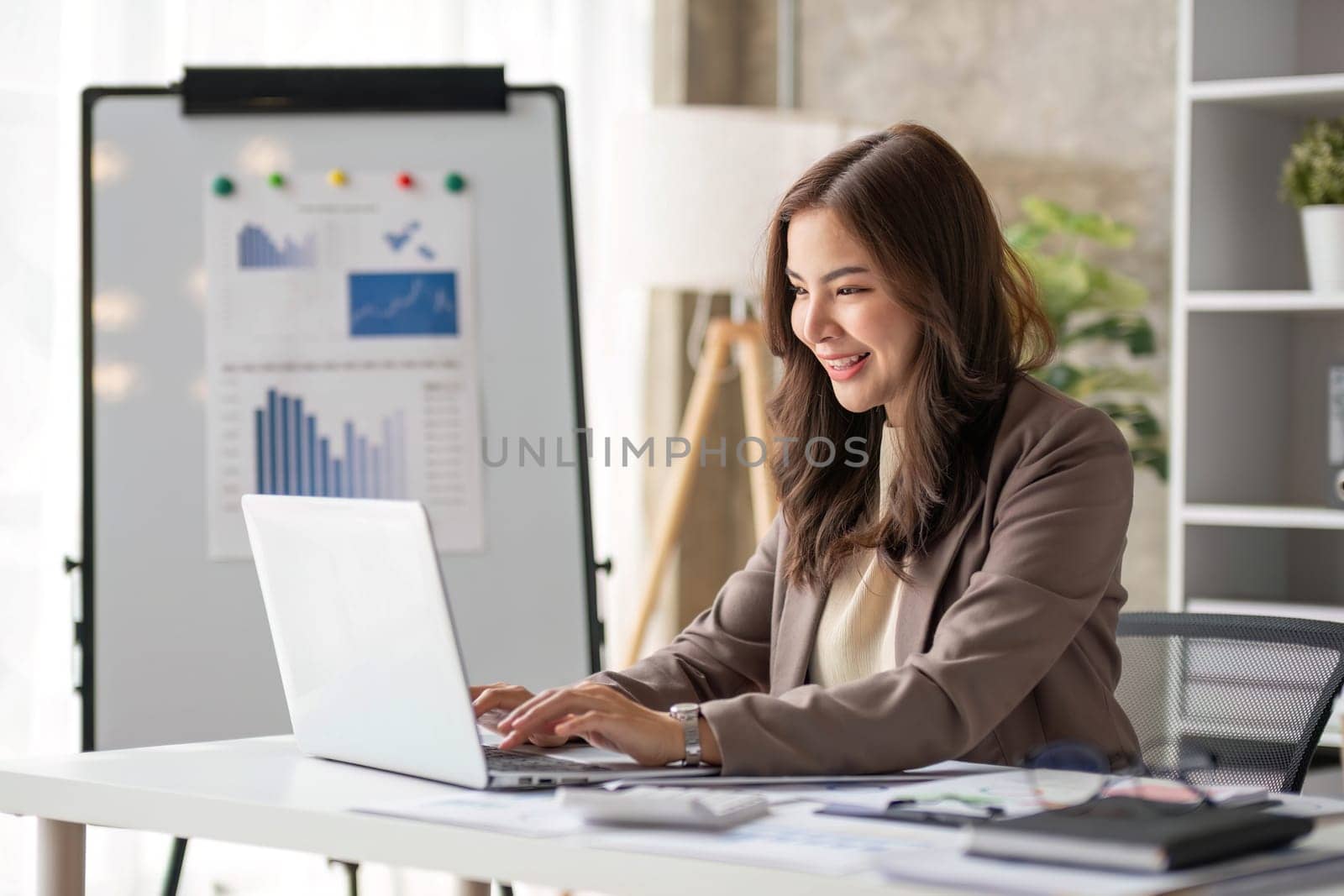 Cheerful business lady working on laptop in office, Asian happy beautiful businesswoman in formal suit work in workplace. Attractive female employee office worker smile...