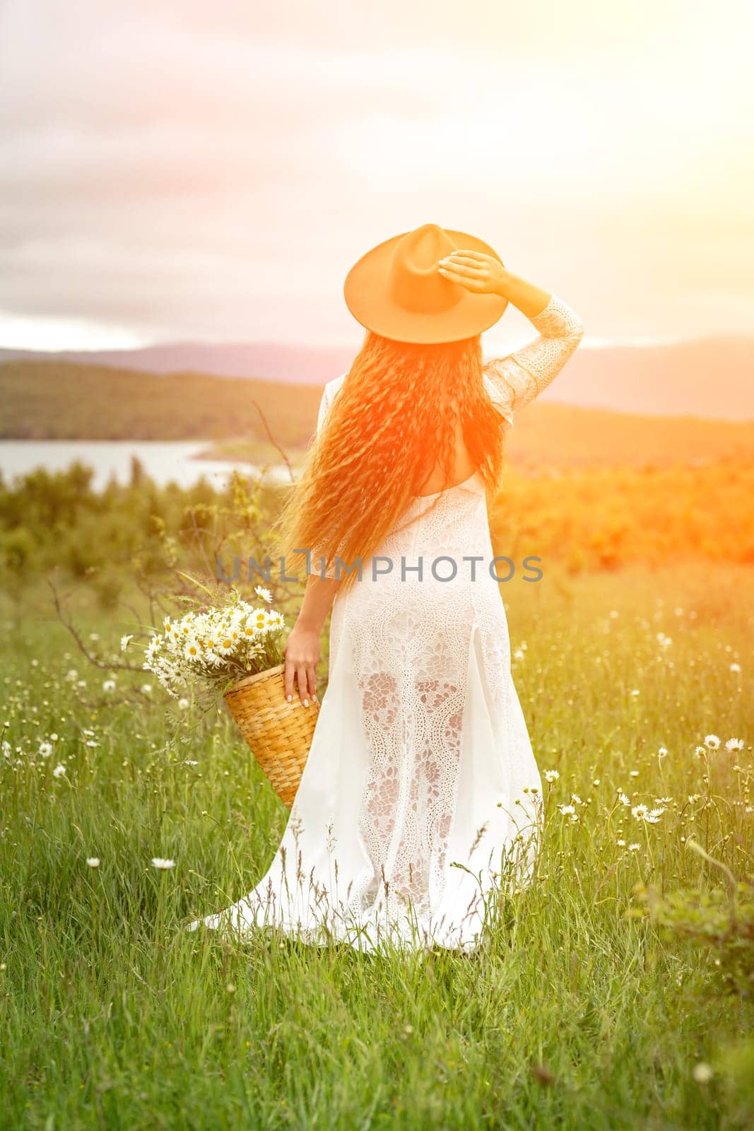 A middle-aged woman in a white dress and brown hat stands with her back on a green field and holds a basket in her hands with a large bouquet of daisies. In the background there are mountains and a lake. by Matiunina