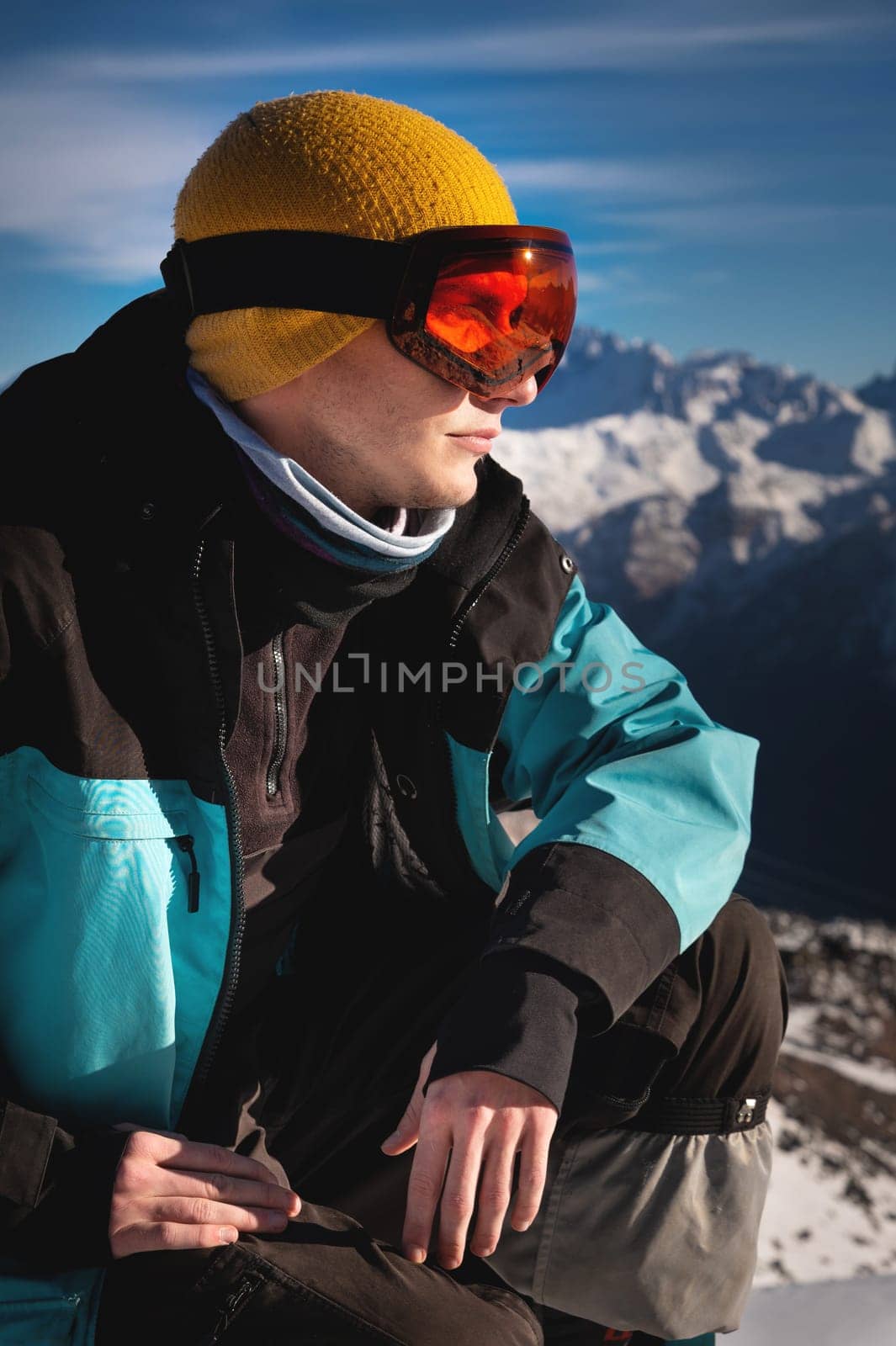 Ski Man sitting on top of a hill. Young man squatting in warm clothes on a sunny day in the snowy mountains by yanik88