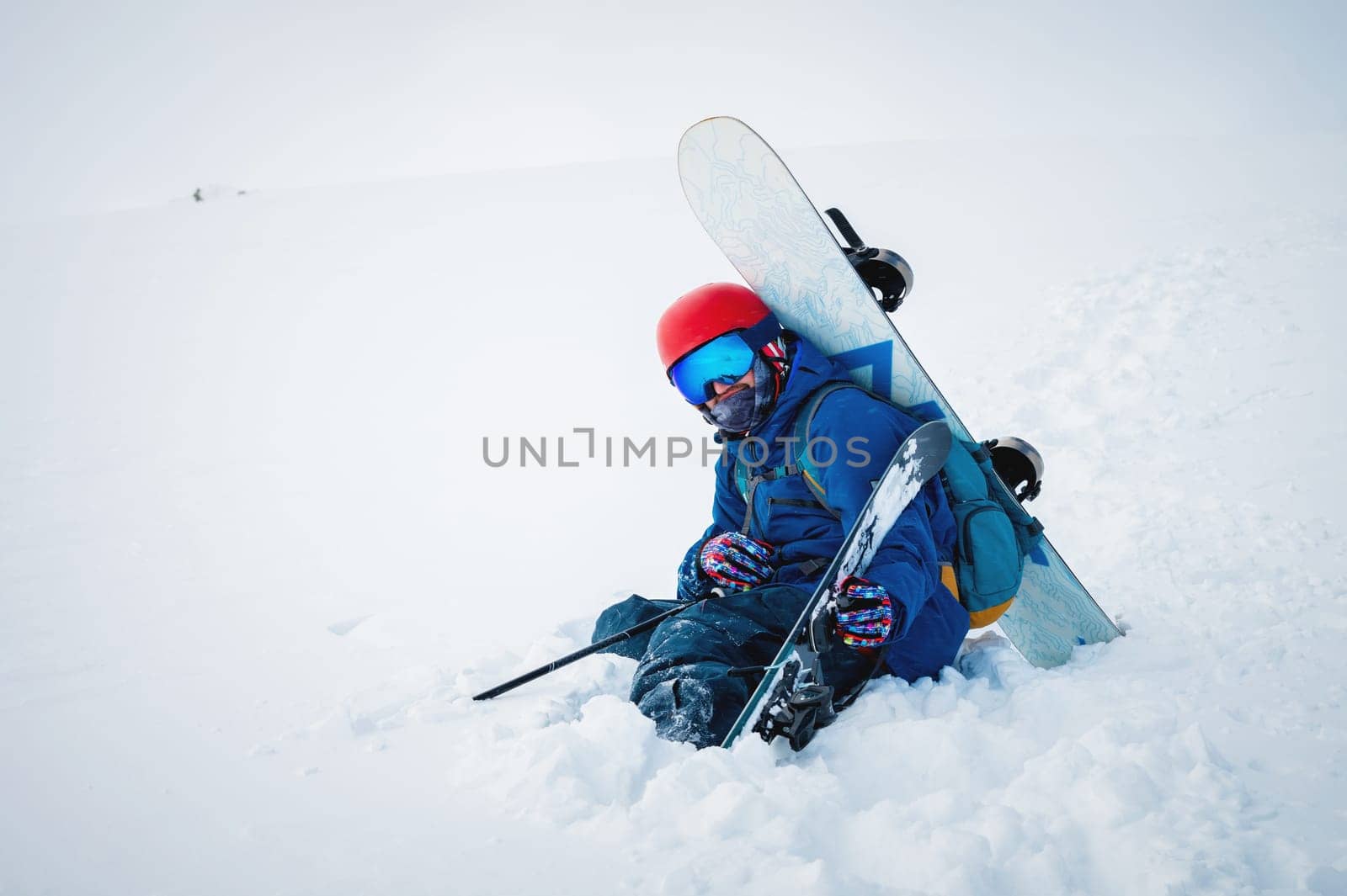 man with ski equipment and a snowboard is sitting a snowy mountain. Blue sky and snowy mountain in the background. by yanik88