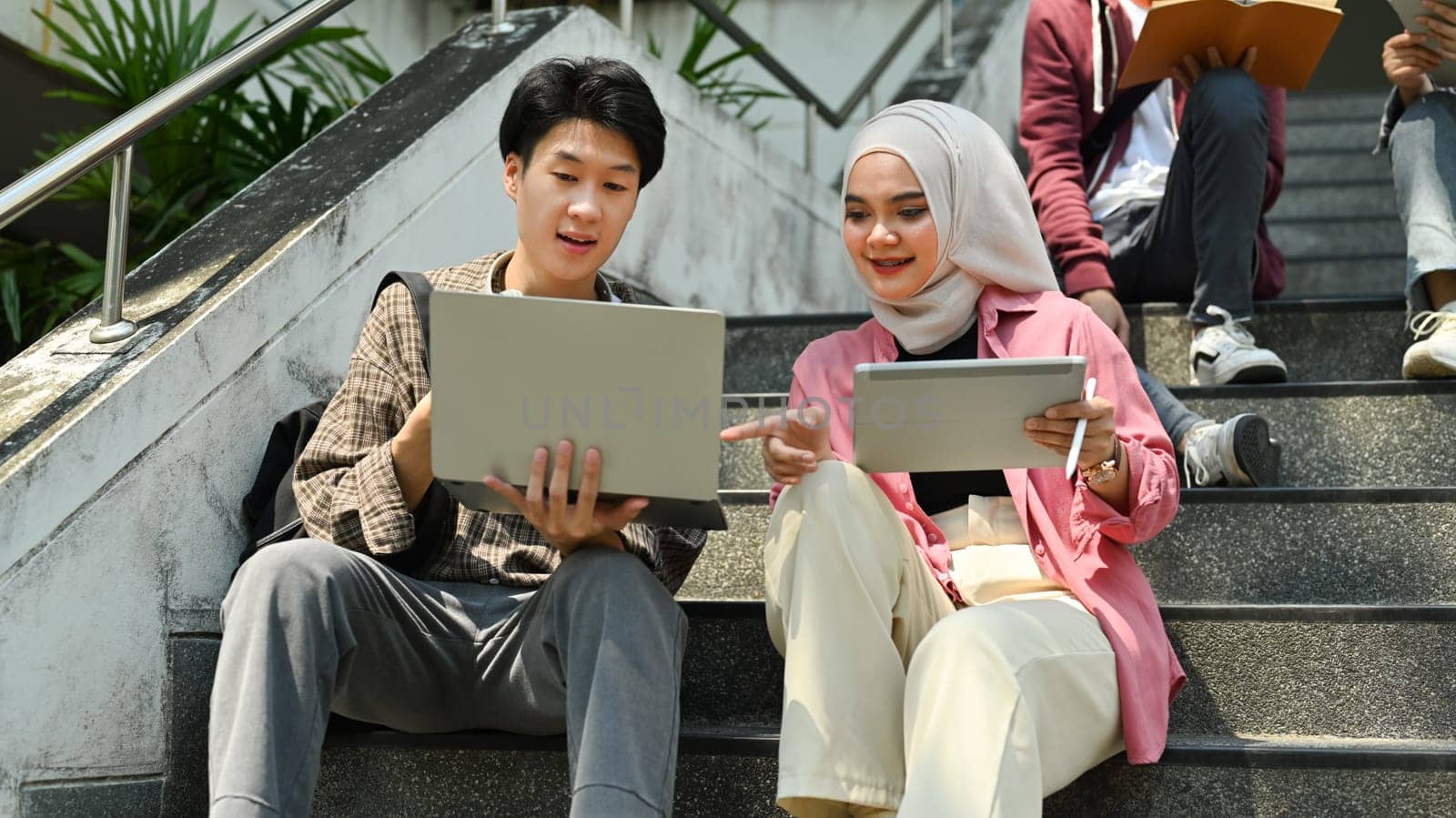 Smiling Asian muslim student doing group project, preparing presentation on laptop with her classmate.