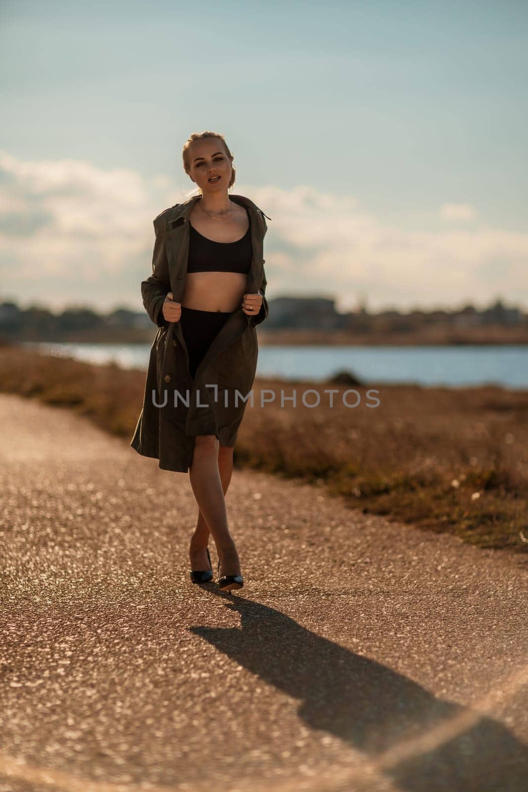 Portrait blonde sea cape. A calm young blonde in an unbuttoned khaki raincoat walks along the seashore, under a raincoat a black skirt and top.