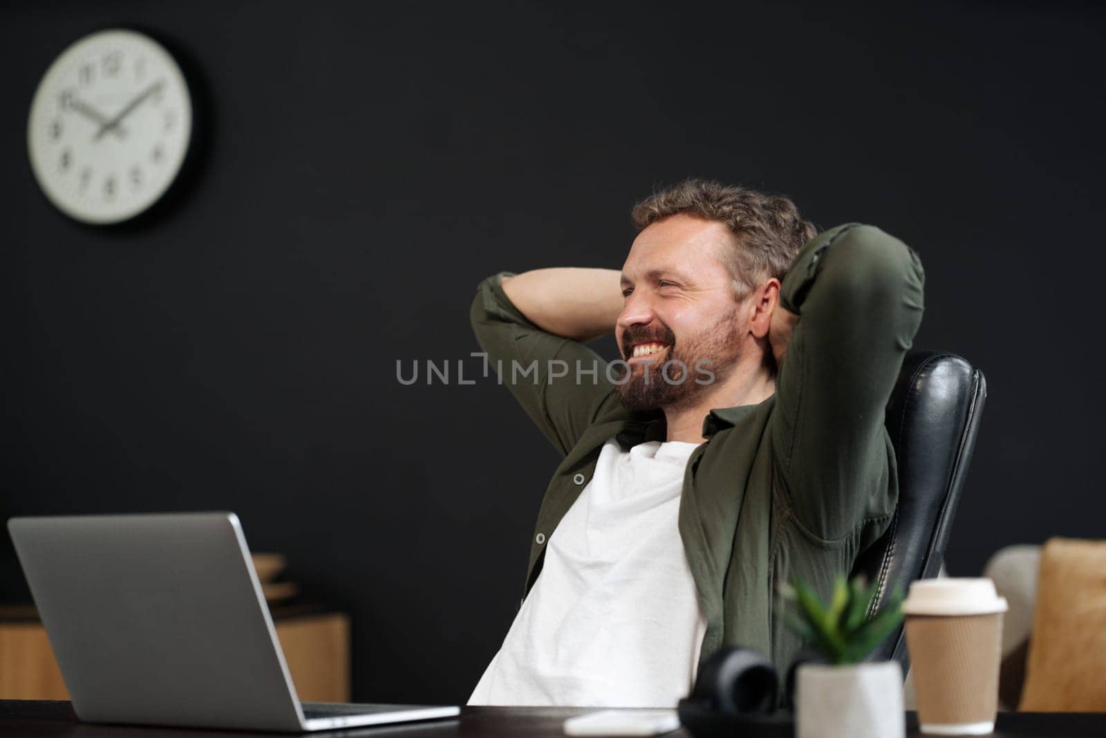 Happy and smiling man taking break and relaxing in office chair. He working on laptop, while enjoying cup of coffee. Modern and productive workplace, with a focus on technology and communication. High quality photo