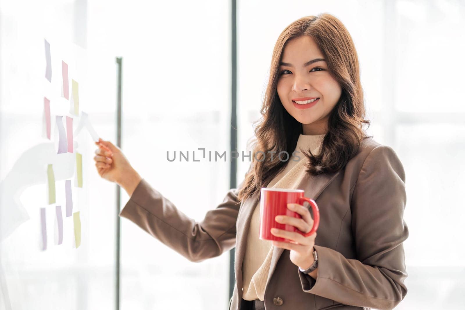 Young smiley attractive, businesswoman using sticky notes in glass wall to writing strategy business plan to development grow to success..