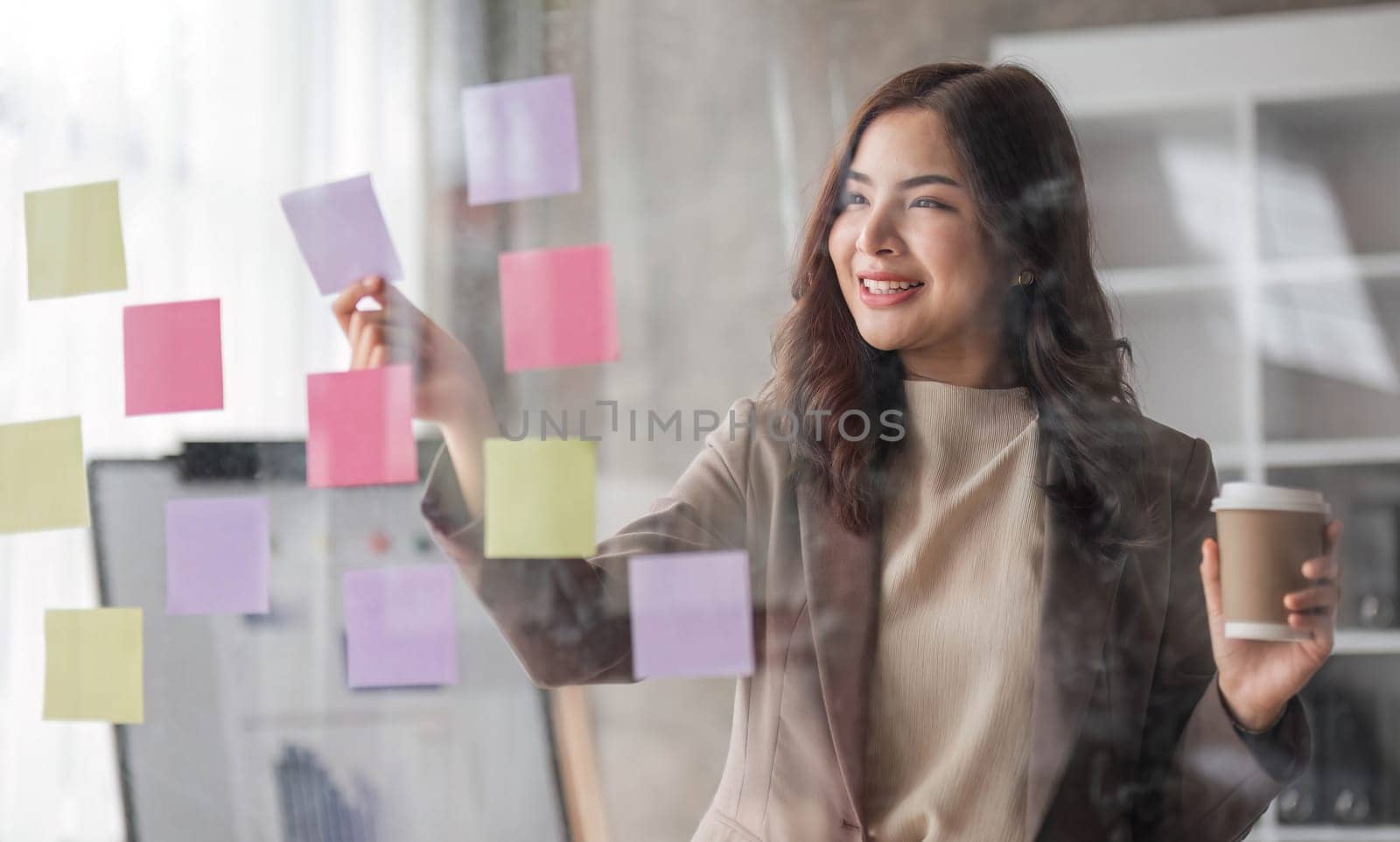 Young smiley attractive, businesswoman using sticky notes in glass wall to writing strategy business plan to development grow to success..
