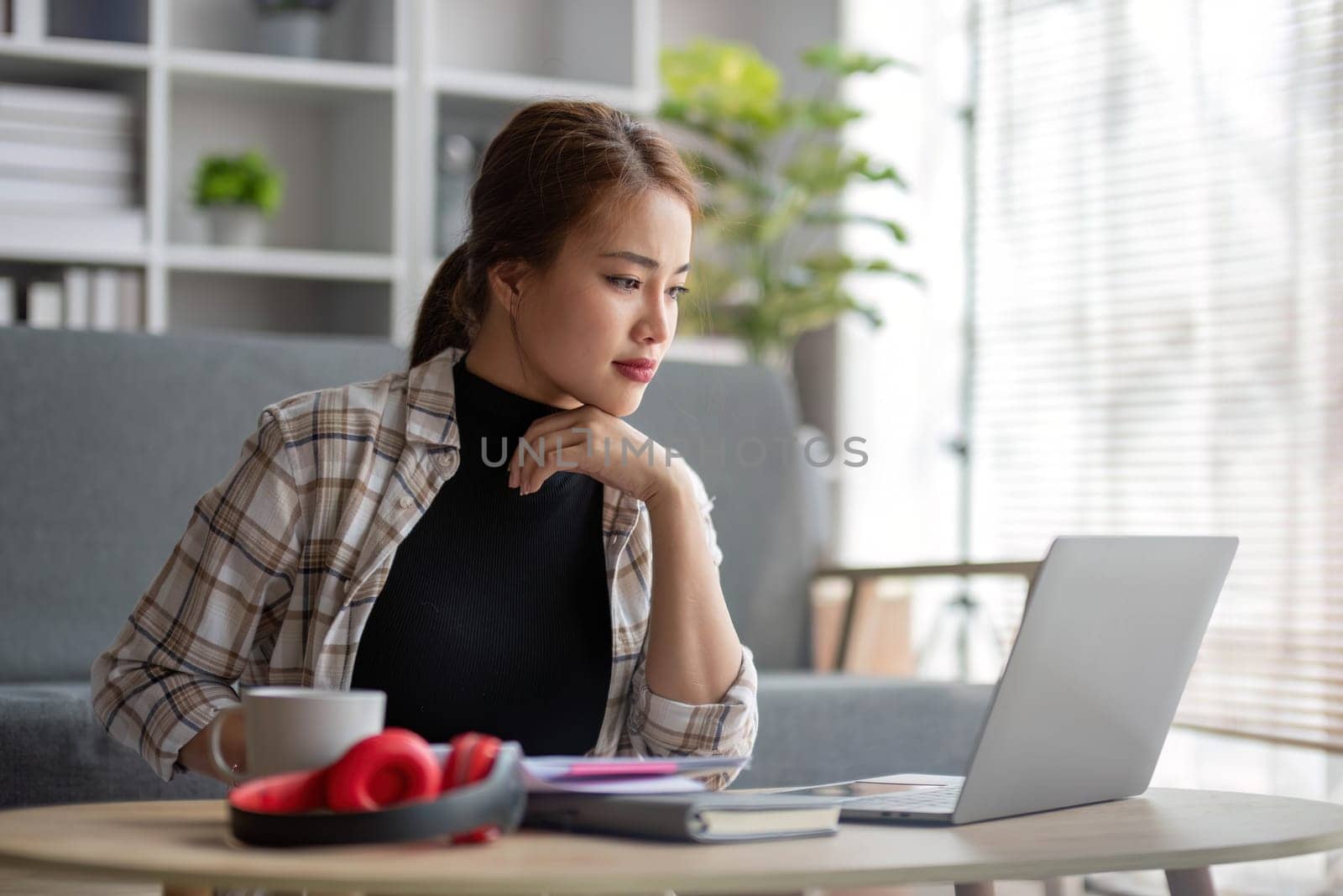 Beautiful and inspired Asian woman working on her tasks on laptop in a living room on the weekend...