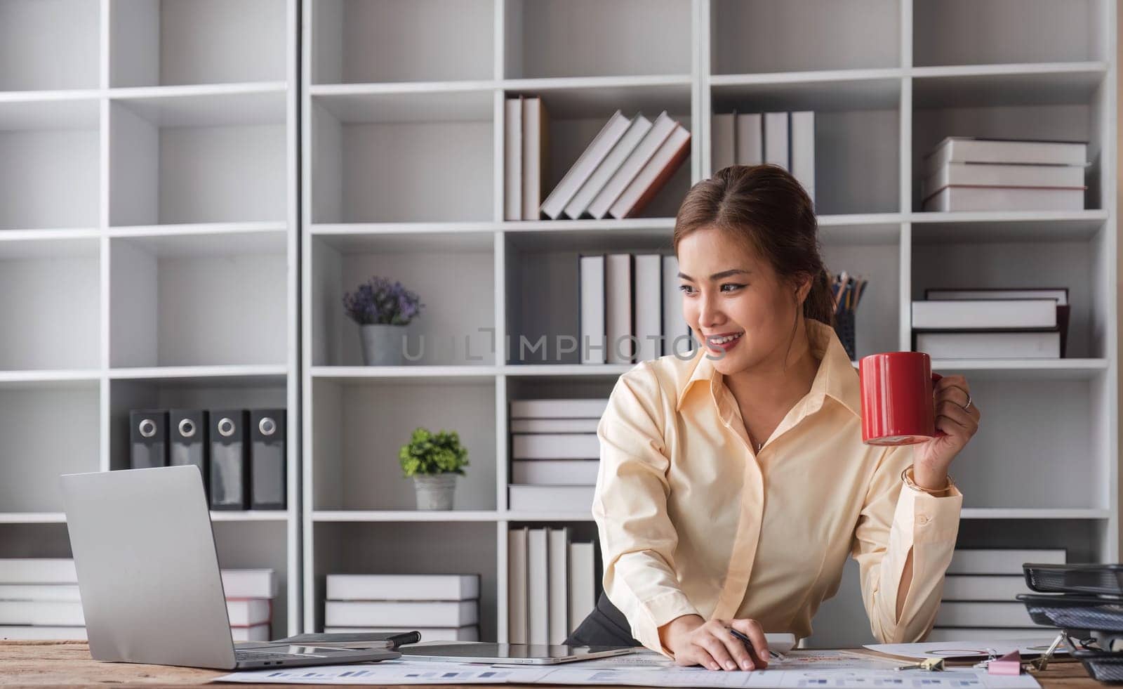 Beautiful and happy young Asian businesswoman looking at her laptop screen while enjoying her morning coffee at her desk in the office...