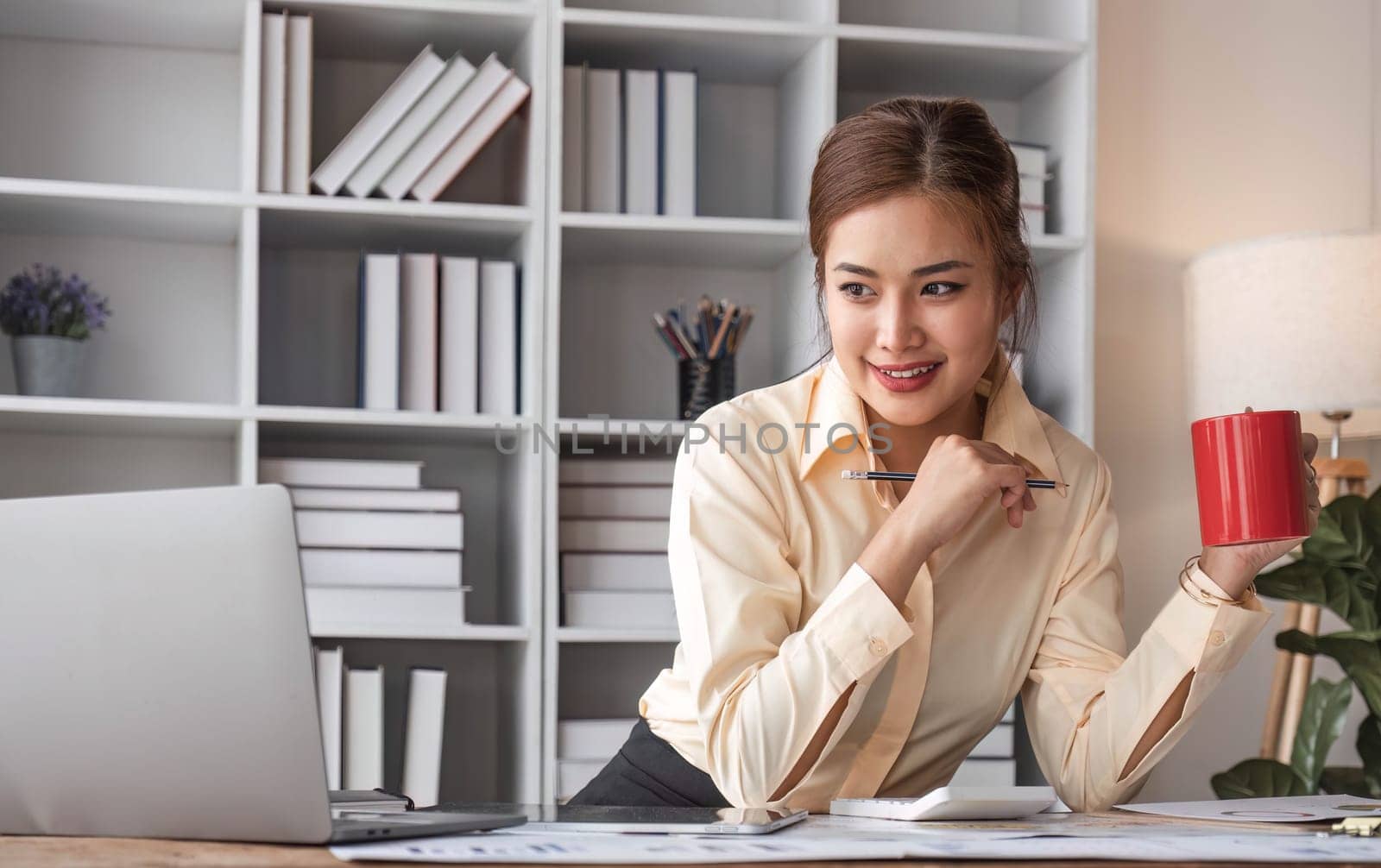 Beautiful and happy young Asian businesswoman looking at her laptop screen while enjoying her morning coffee at her desk in the office...