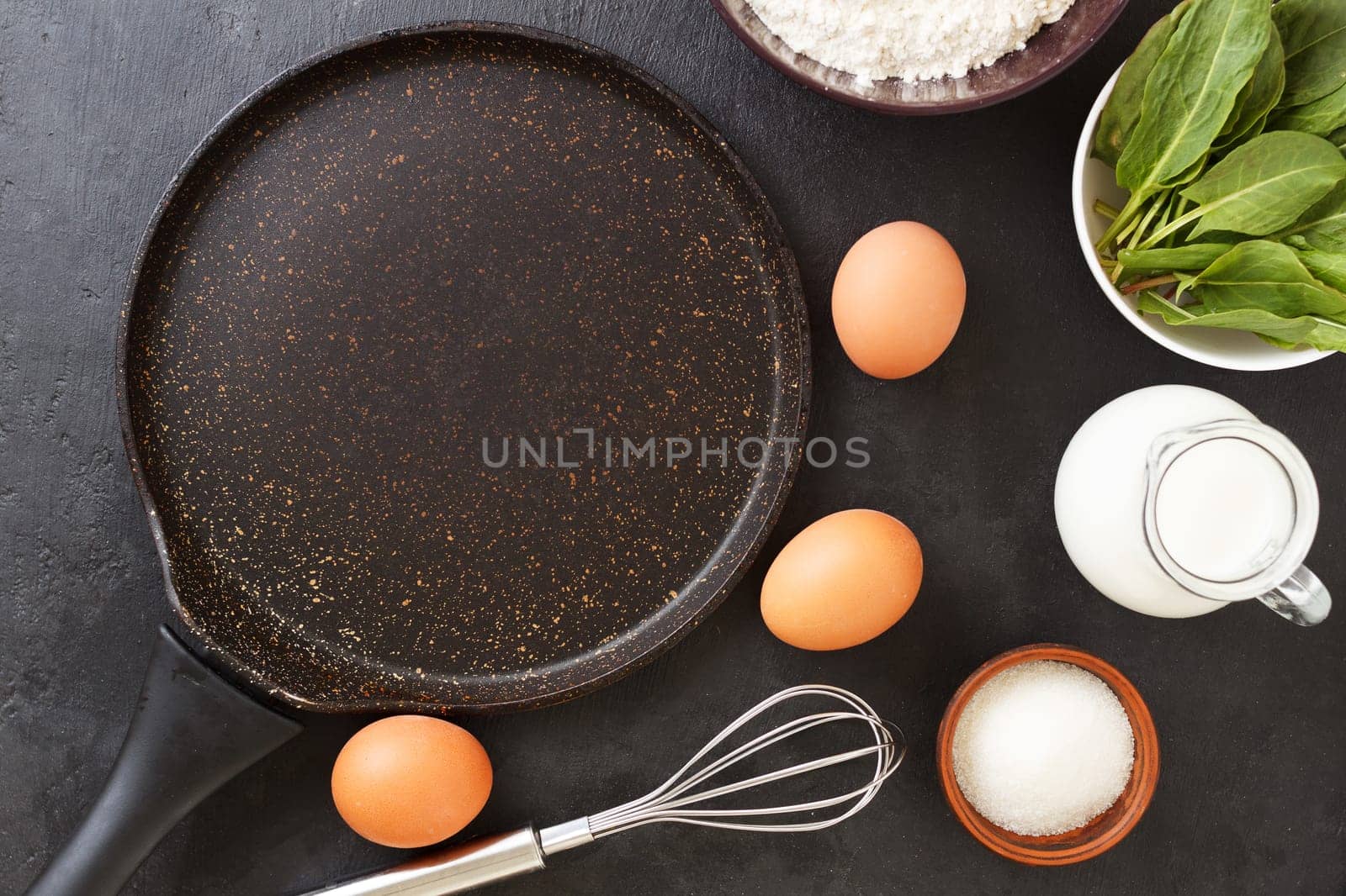 An empty pan for making spinach pancakes on a black background with ingredients: eggs, spinach, milk, flour. by lara29