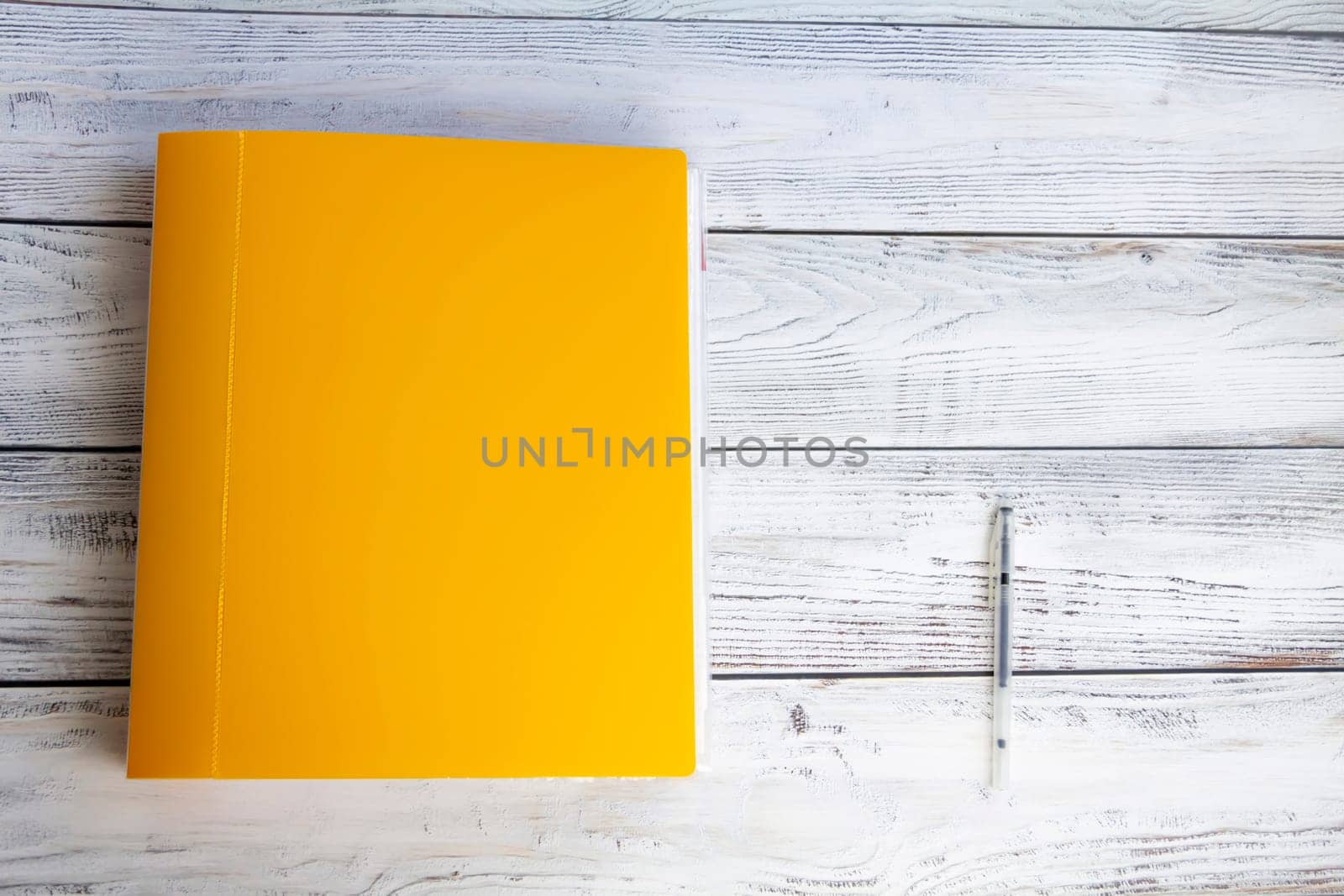 A bright, yellow, thick, stationery folder with a large number of documents and a ballpoint pen for office work and university studies on a light, wooden background.