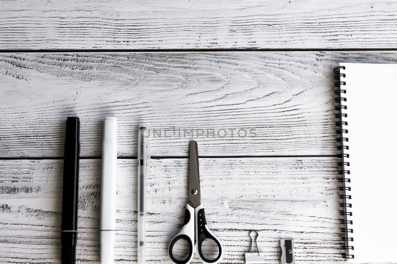 A sketchbook with white sheets, a clip, a metal sharpener, a black and white marker, a writing pen and scissors in black and white tones and shades on wooden background.