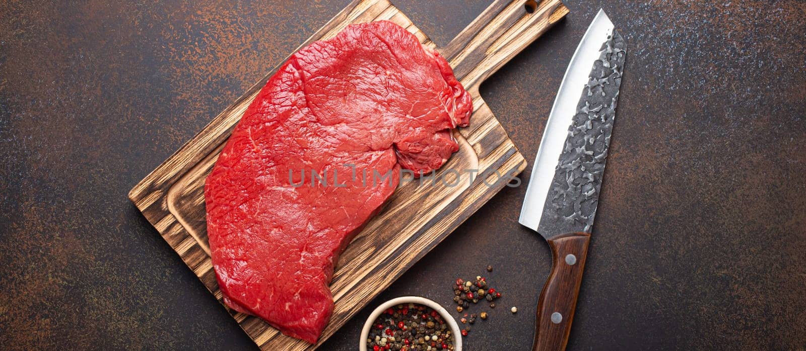 Raw uncooked top round beef steak on wooden cutting board with big kitchen knife and pepper on dark brown rustic stone background top view, cooking meat steak concept