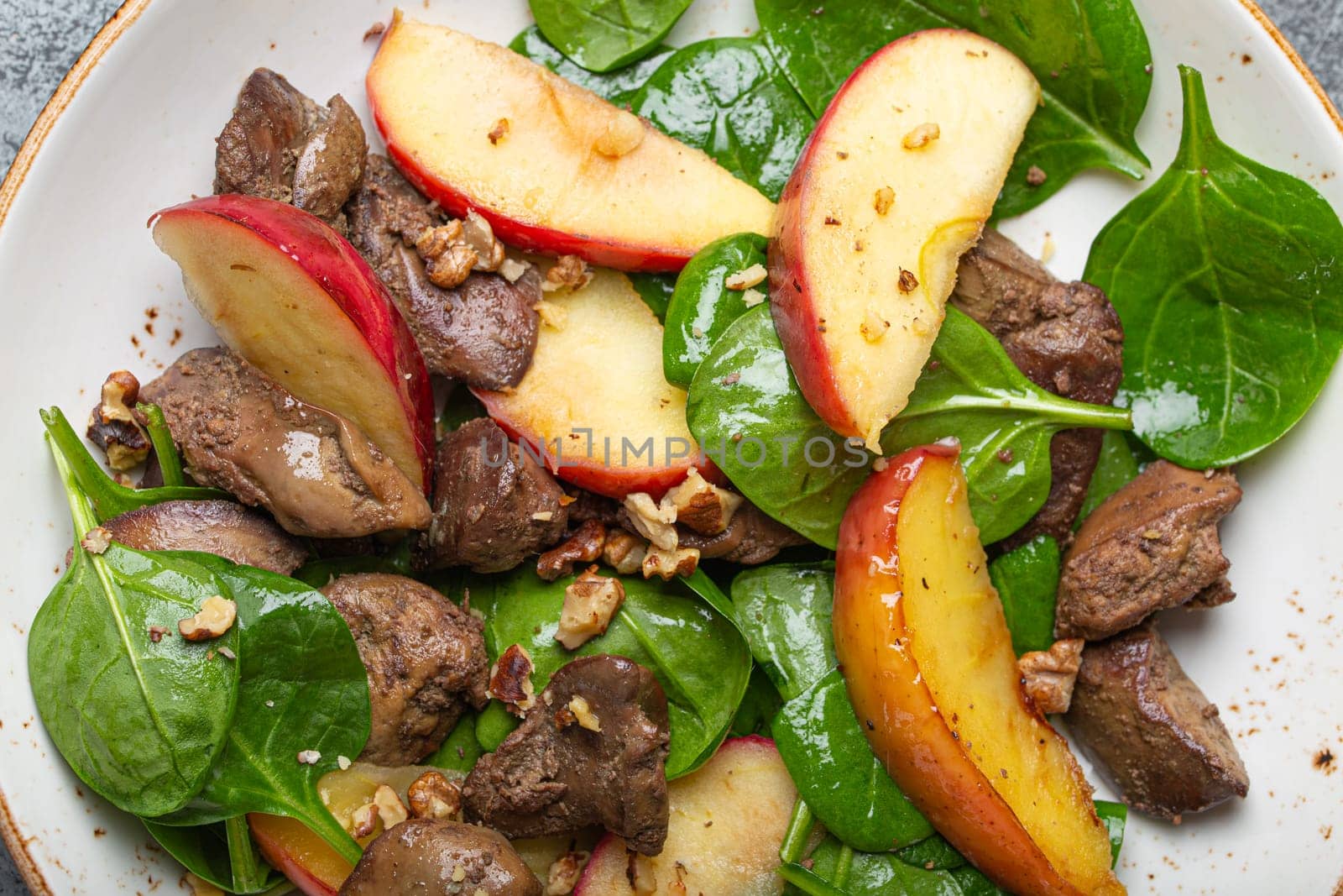 Close Up of Healthy Salad with Iron Rich Ingredients Chicken Liver, Apples, Fresh Spinach and Walnuts on White Ceramic Plate Top View.
