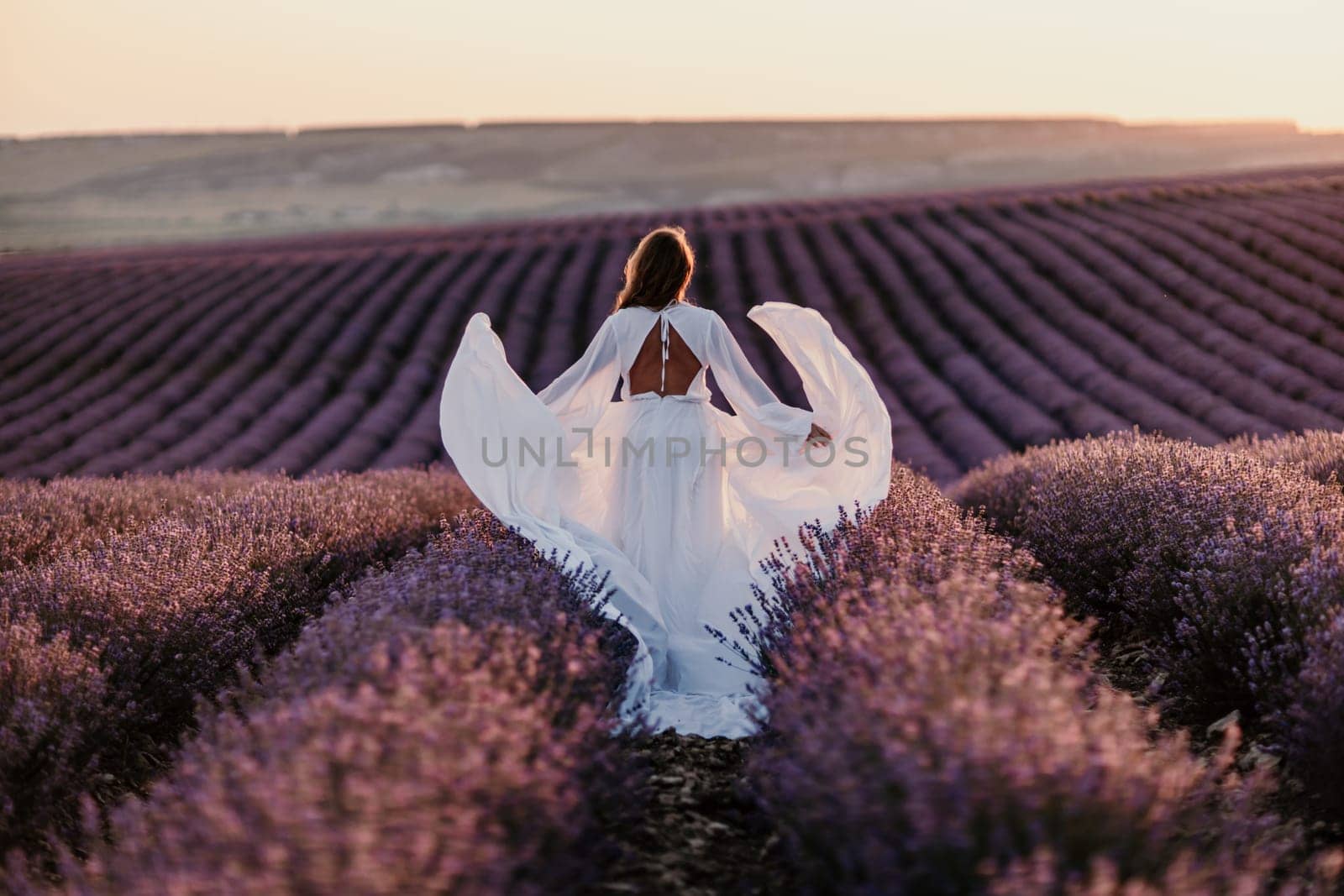 Happy woman in a white dress and straw hat strolling through a lavender field at sunrise, taking in the tranquil atmosphere