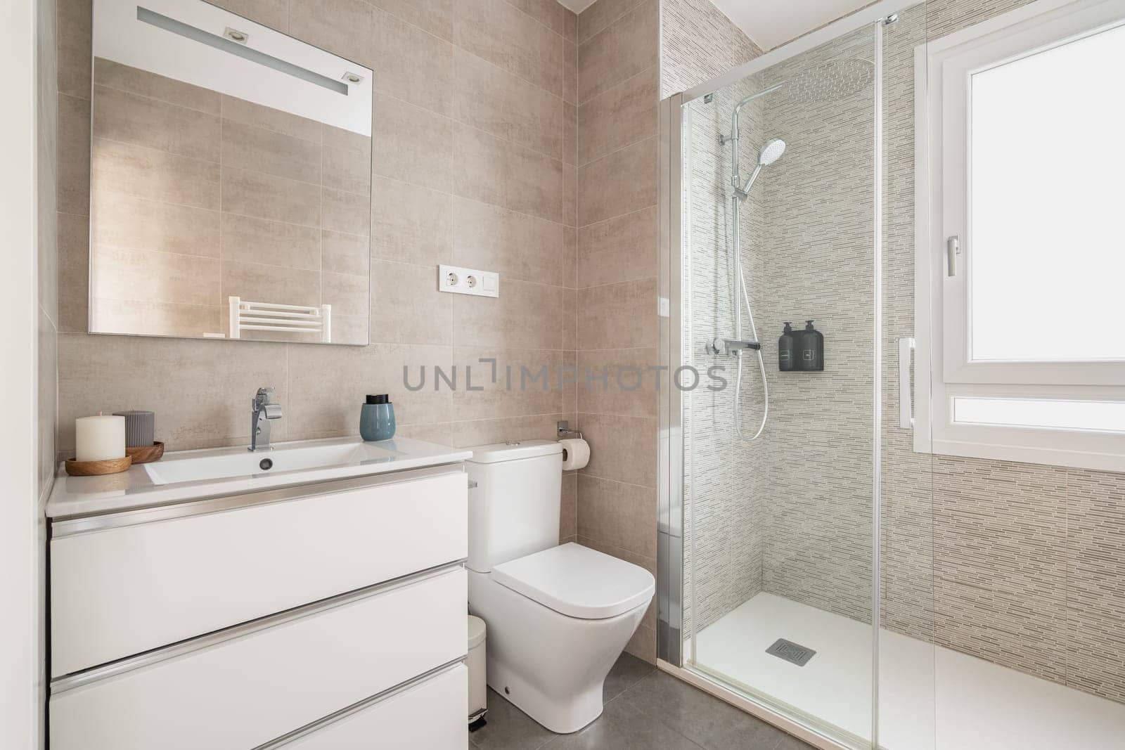 Beige tiled bathroom with glass shower doors with toilet and sink over cupboard with mirror and daylight window. Modern bathroom design concept.