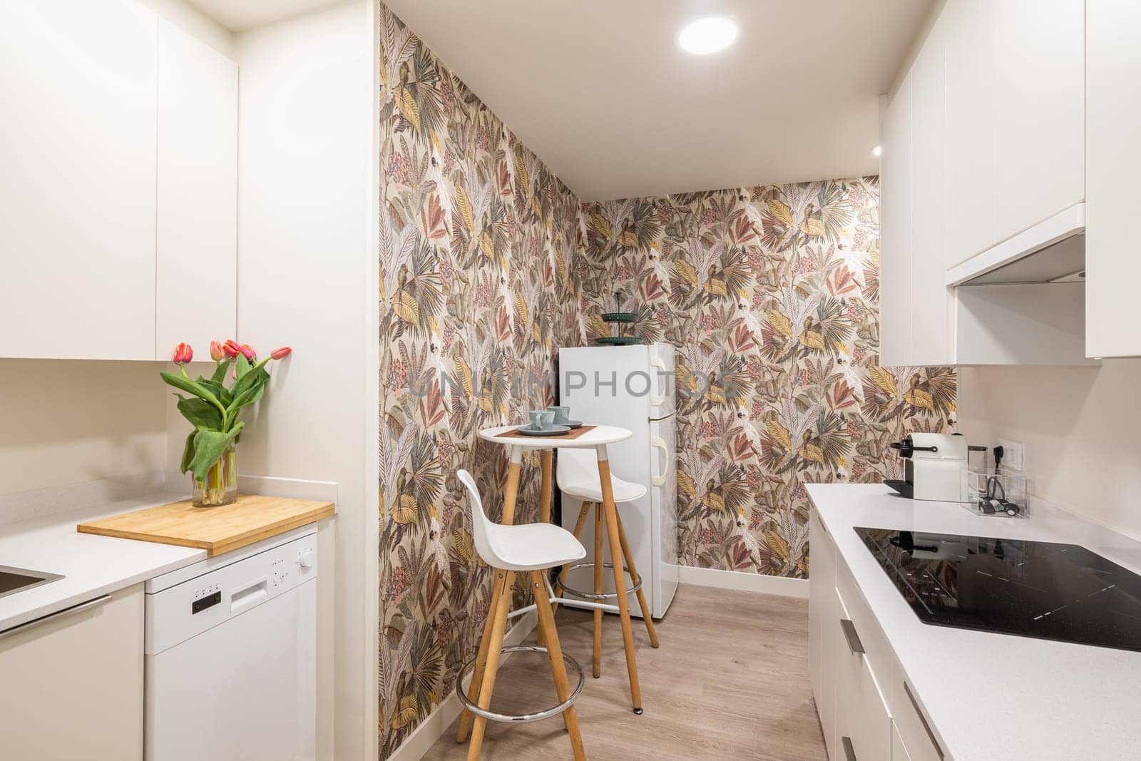 Compact kitchen with a small table and a comfortable cooking surface with modern appliances and wall cabinets with floral wallpaper. Kitchen concept after renovation in small apartments.