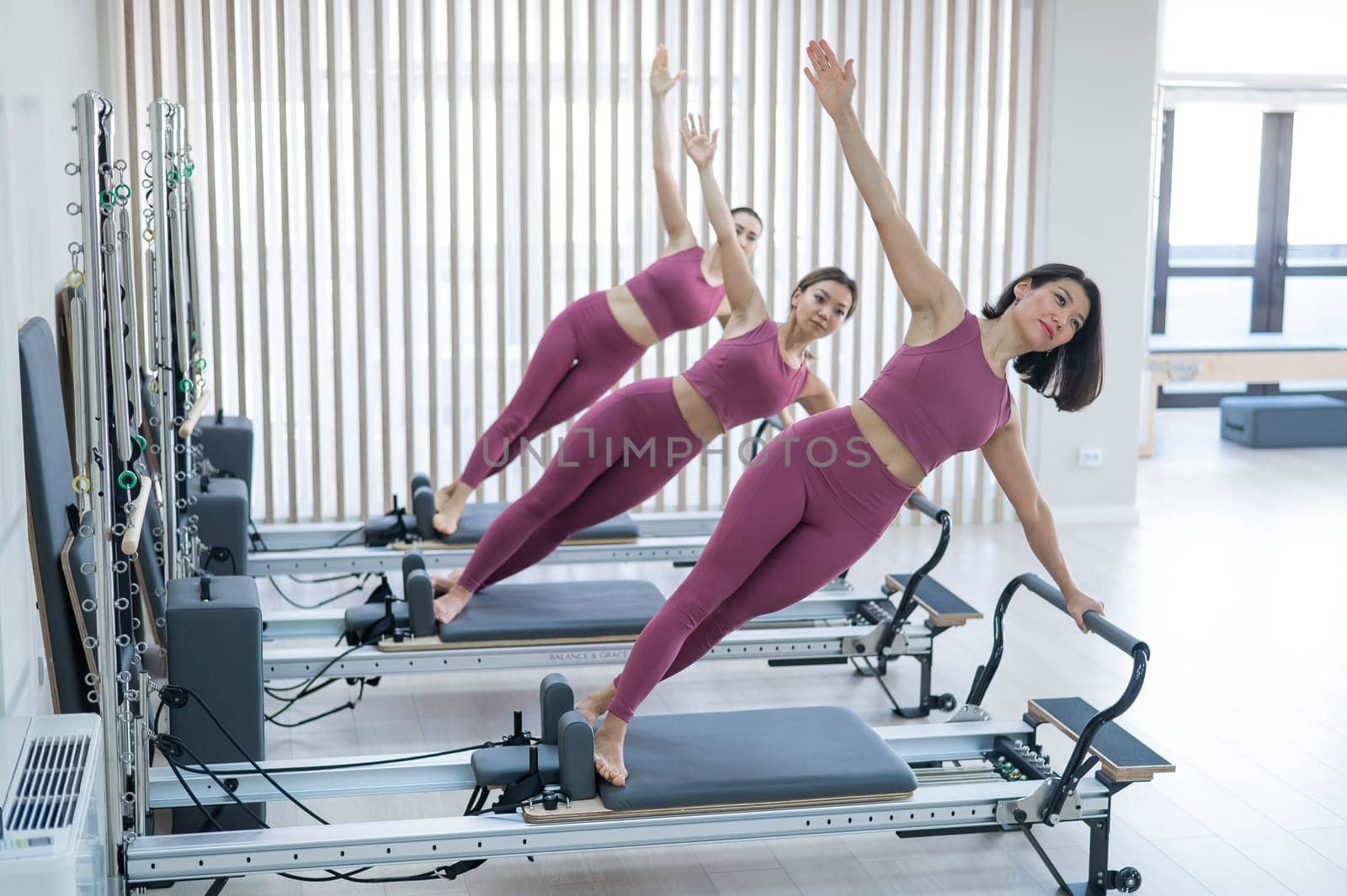Three Asian women in pink sportswear doing side planks on a reformer. Pilates classes