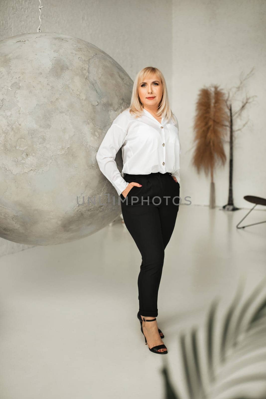 A fashionable woman In a white shirt and black pants poses in the interior.