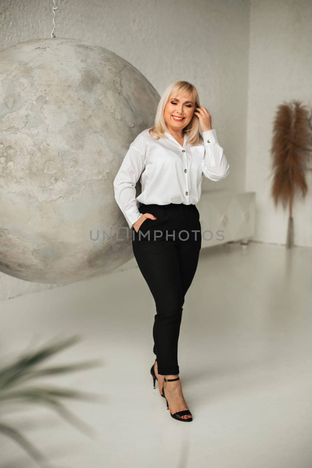 A fashionable woman In a white shirt and black pants poses in the interior.