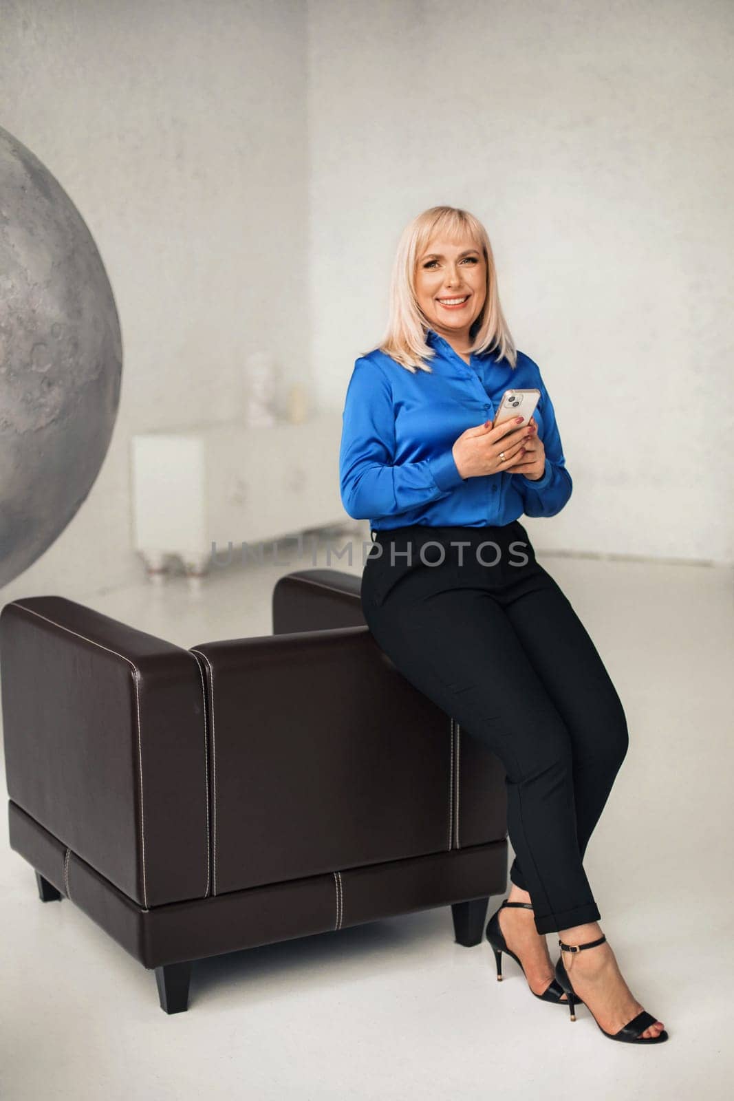 A fashionable woman in a blue shirt and black trousers poses in the interior with a smartphone in her hands.