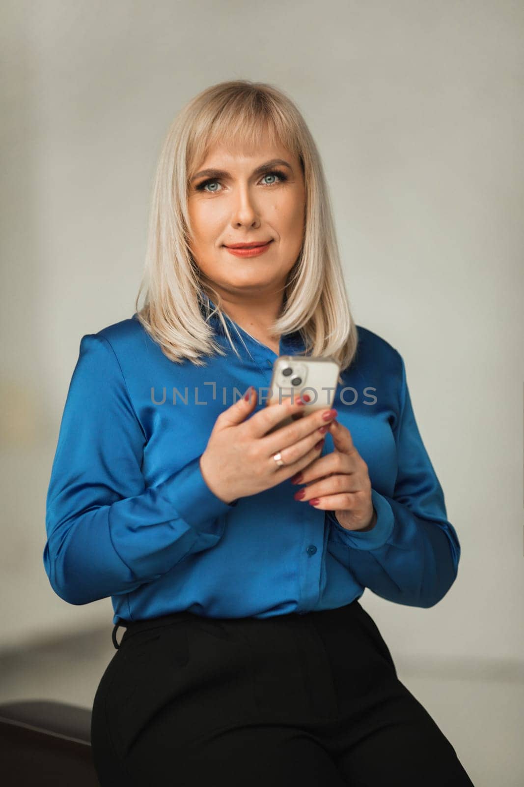 A fashionable woman in a blue shirt and black trousers poses in the interior with a smartphone in her hands.